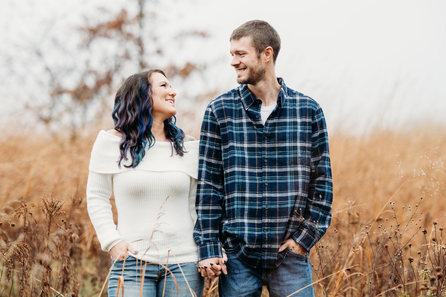 A couple in casual fall attire stands together smiling in a golden autumn field during sunset.