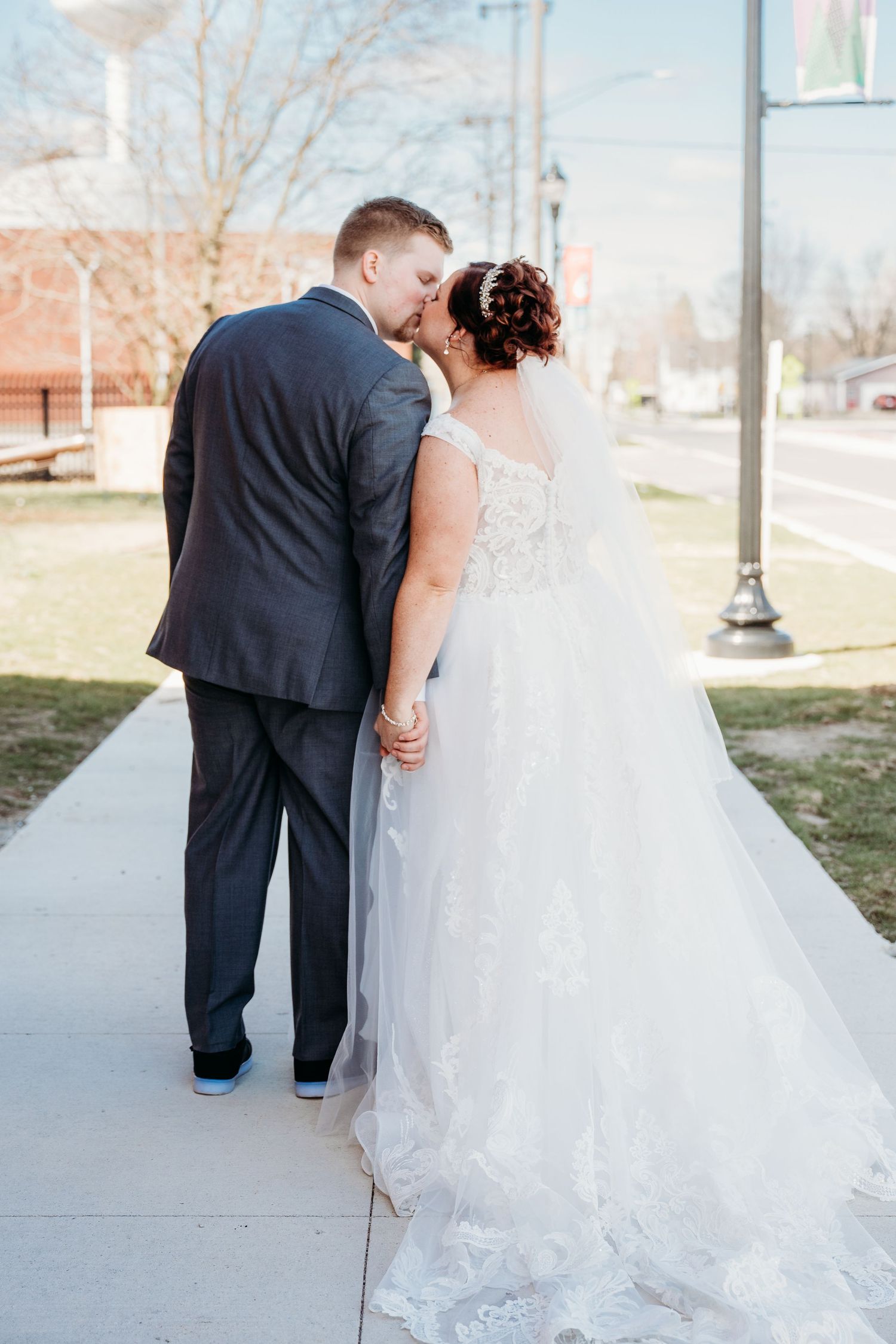 Newlywed couple in formal attire standing on a sidewalk with their backs to the camera.