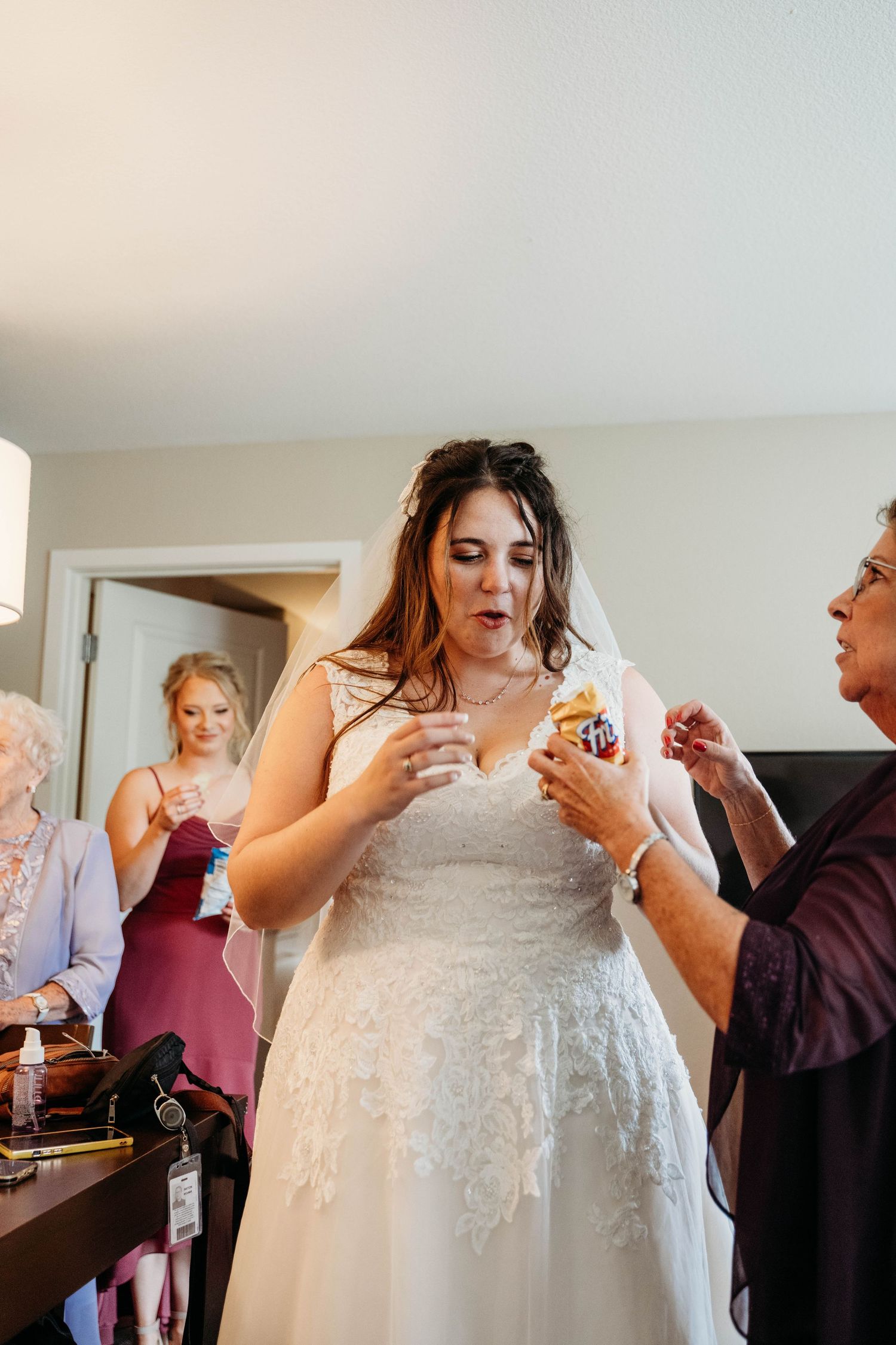 A person in a white lace wedding dress looks down at a small object during preparations with others in the room.