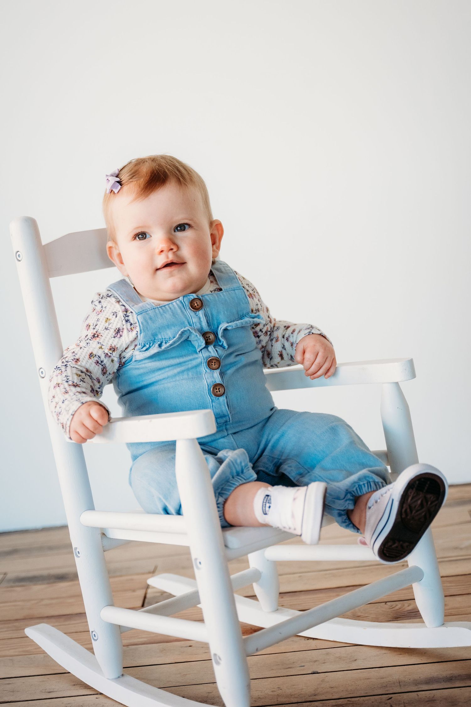 Baby wearing blue overalls and floral shirt sits in white wooden rocking chair on hardwood floor.