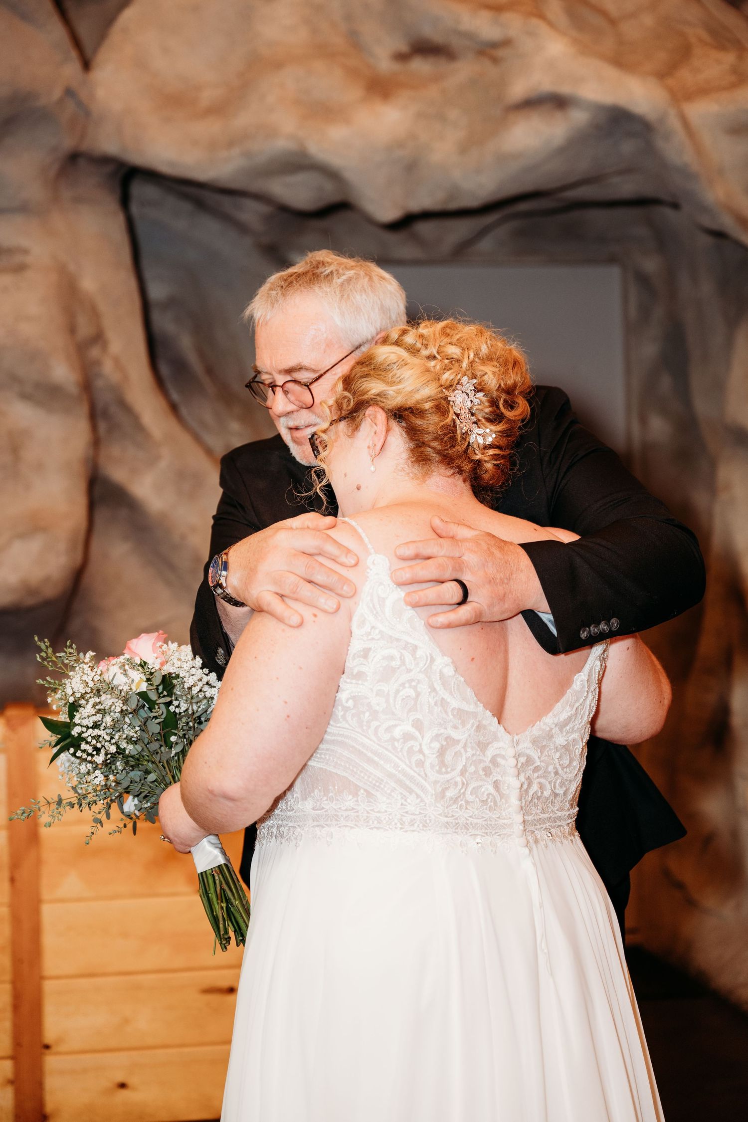 A tender embrace at a wedding between someone in a white lace gown holding a bouquet and another in a black suit.