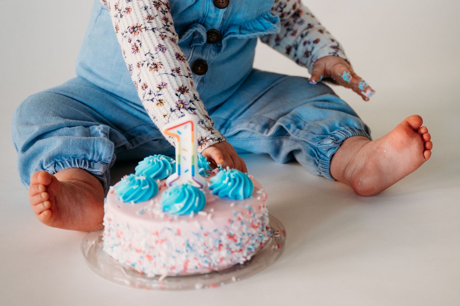 A white birthday cake with blue frosting decorations next to small feet wearing blue clothing.
