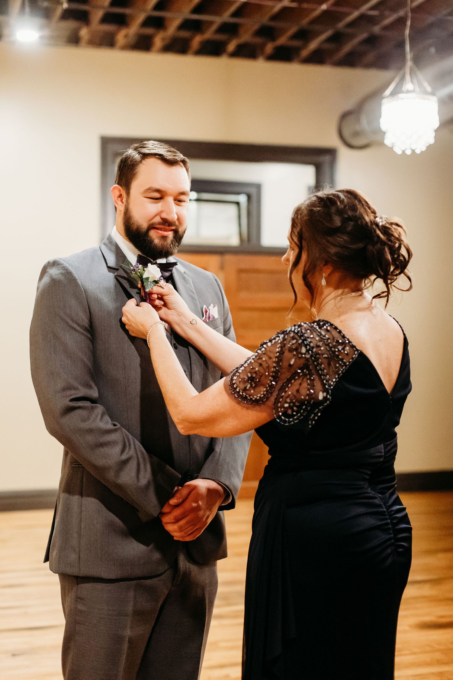 Someone helps adjust a bowtie on a groom wearing a gray suit while wearing a sparkling black evening dress.