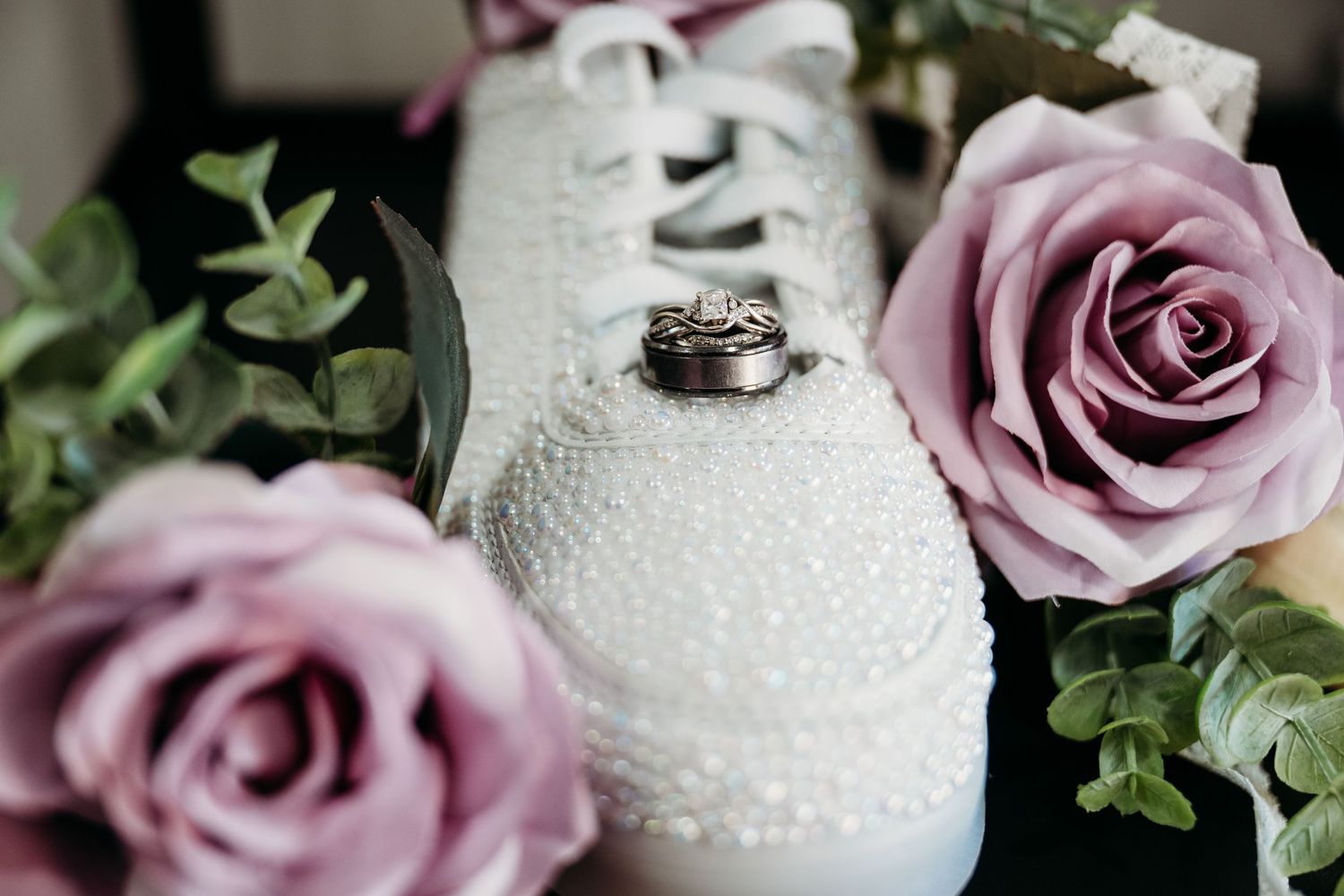 Wedding rings displayed on white bridal shoes surrounded by dusty pink roses and greenery.