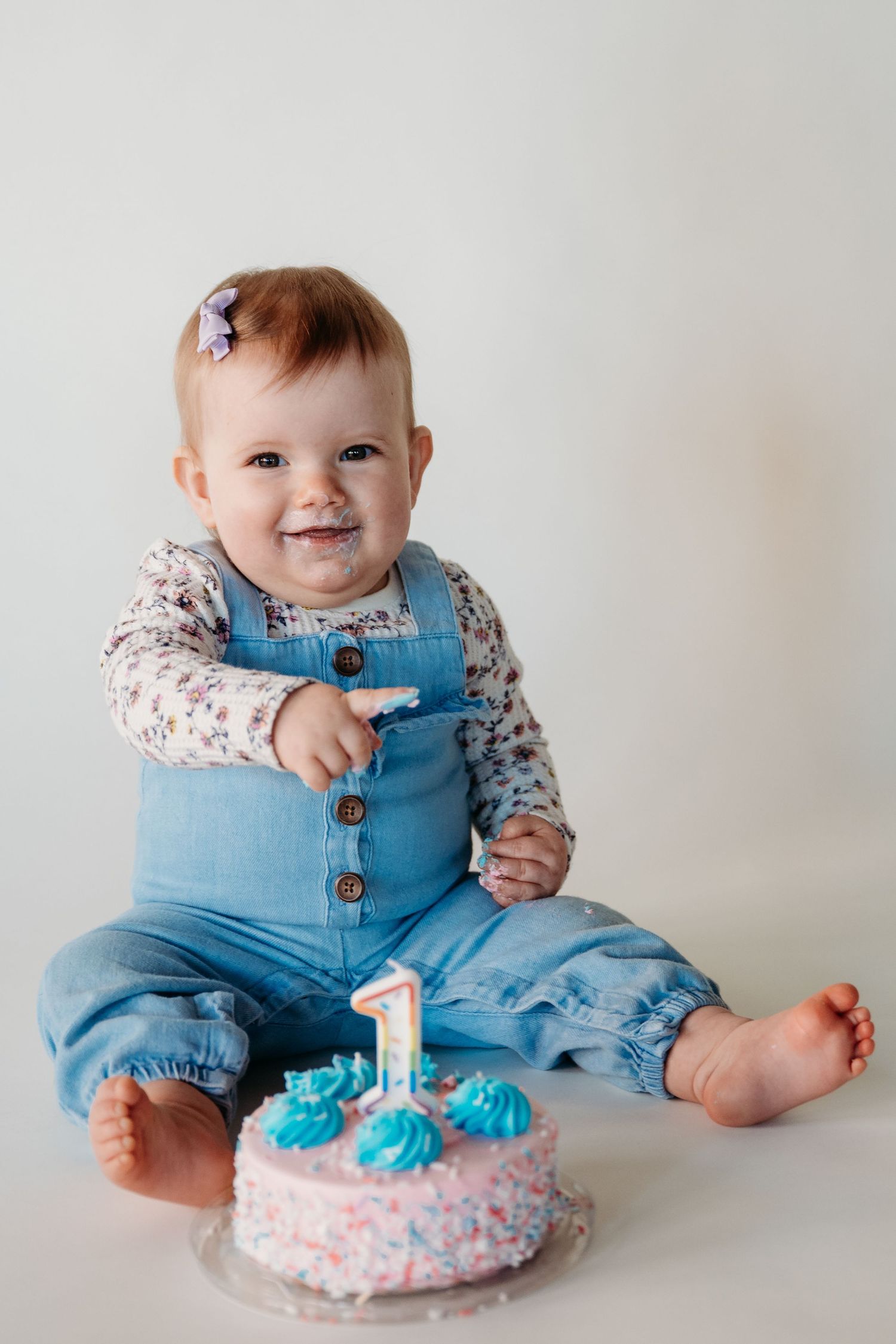 A baby in blue overalls sits beside a pink and blue first birthday cake with a happy expression.