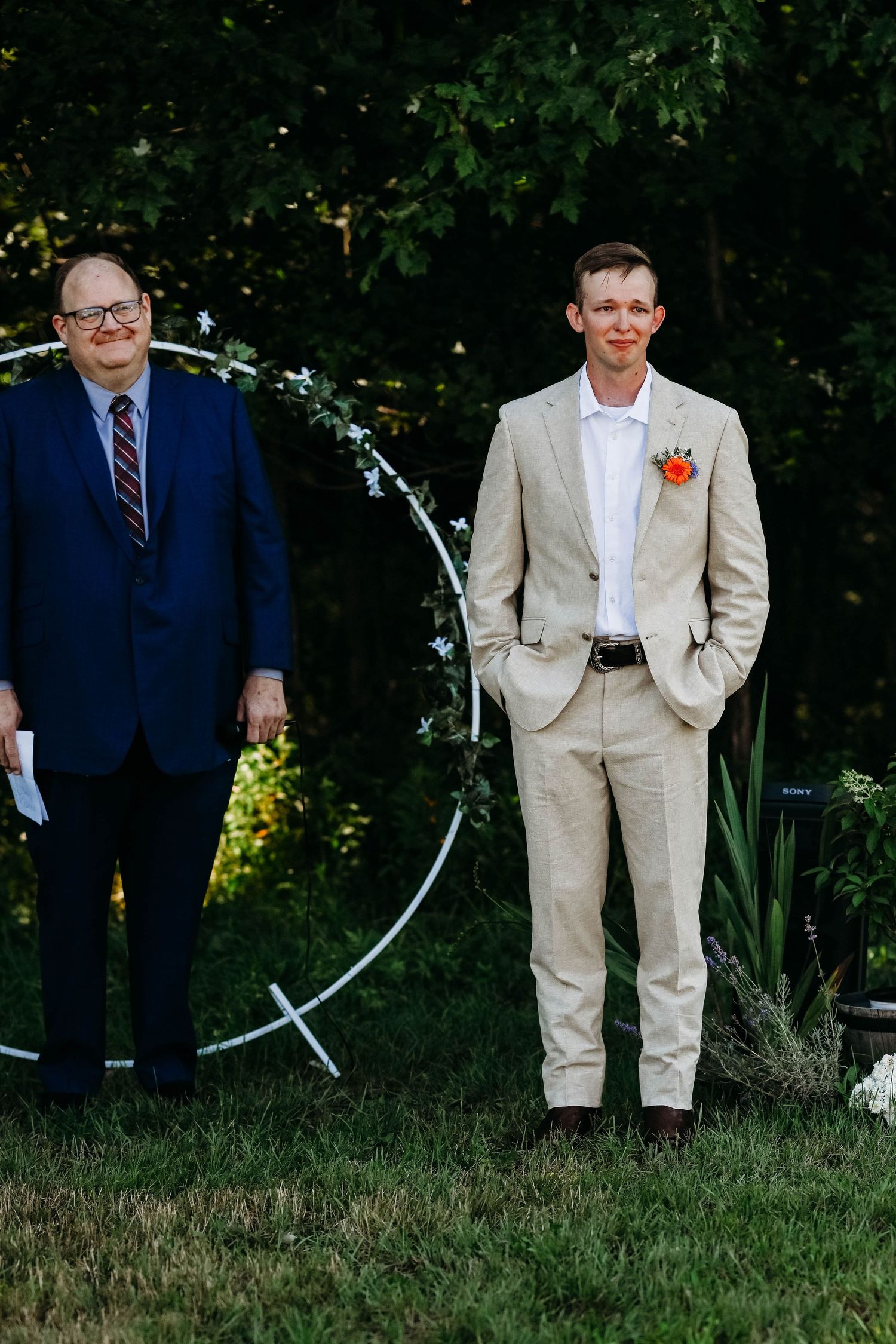 Two people in formal attire stand by a circular wedding arch outdoors with greenery in the background.