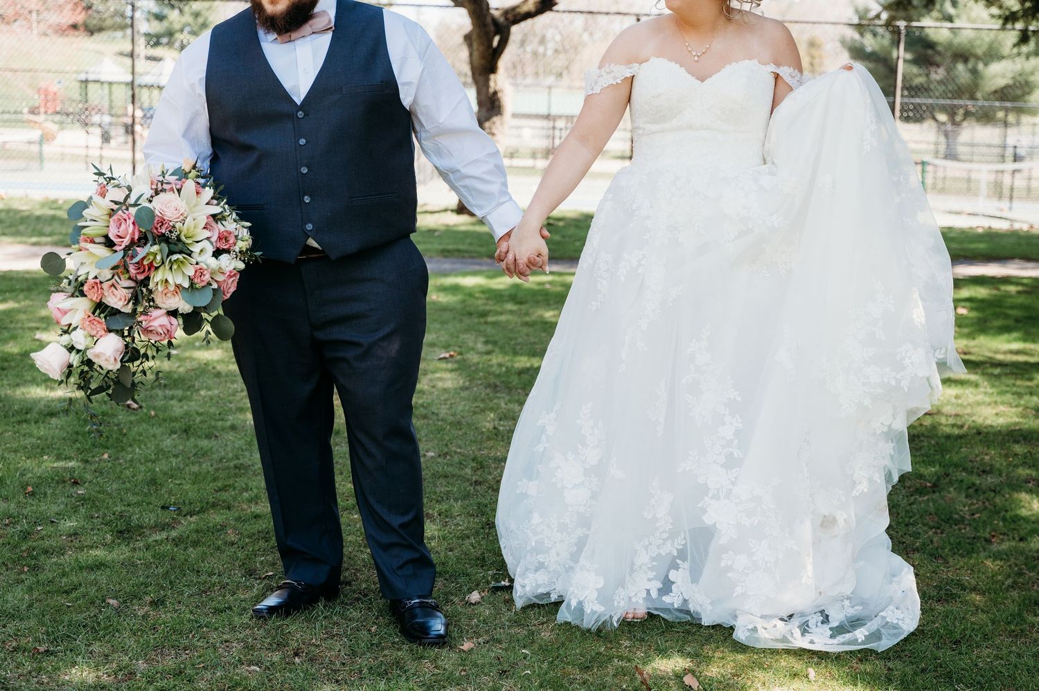 A couple holds hands outdoors during their wedding ceremony with the bride carrying a pink and peach floral bouquet.