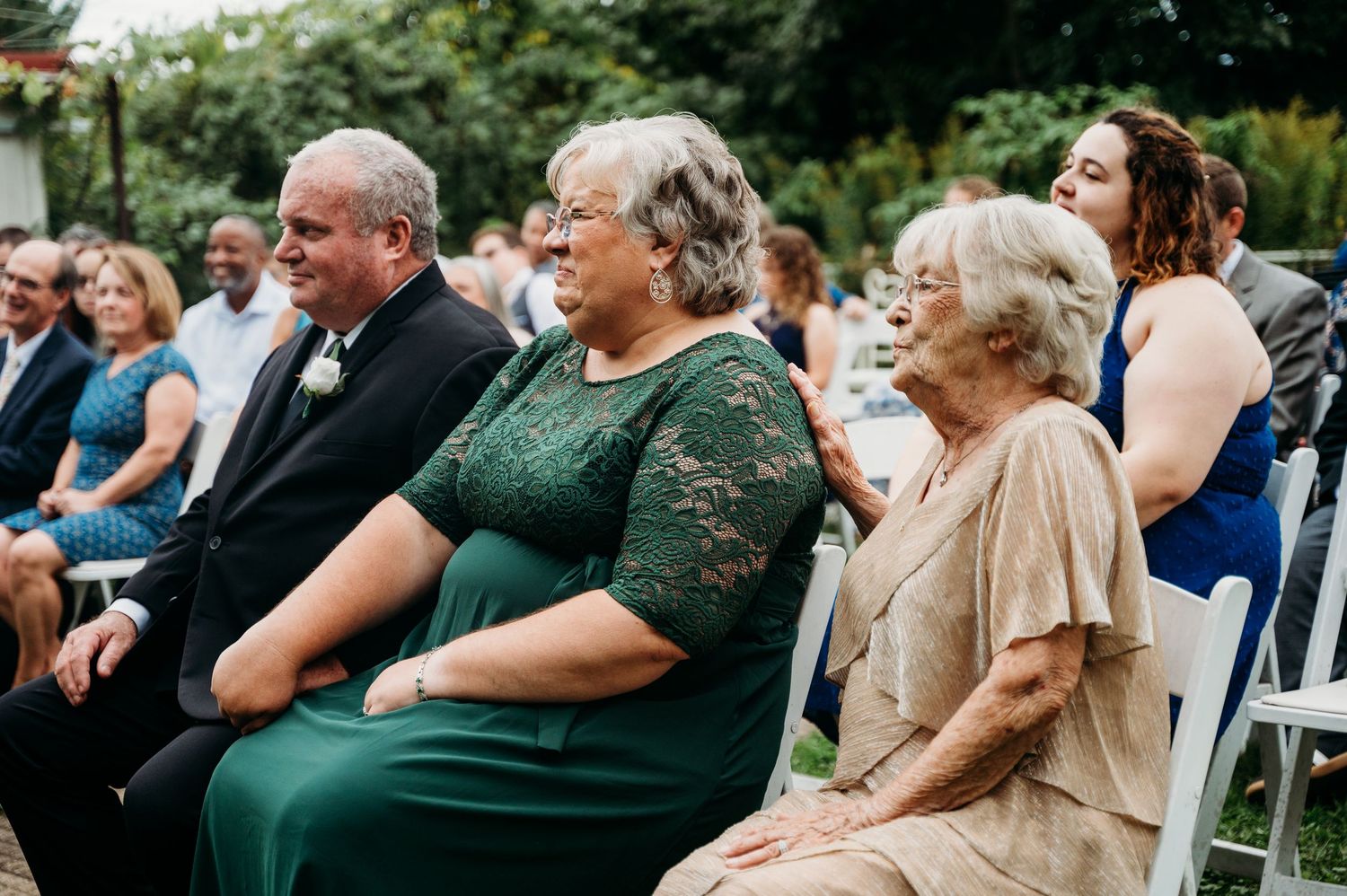 Attendees sitting in white folding chairs during an outdoor wedding ceremony dressed in green and beige formal attire.