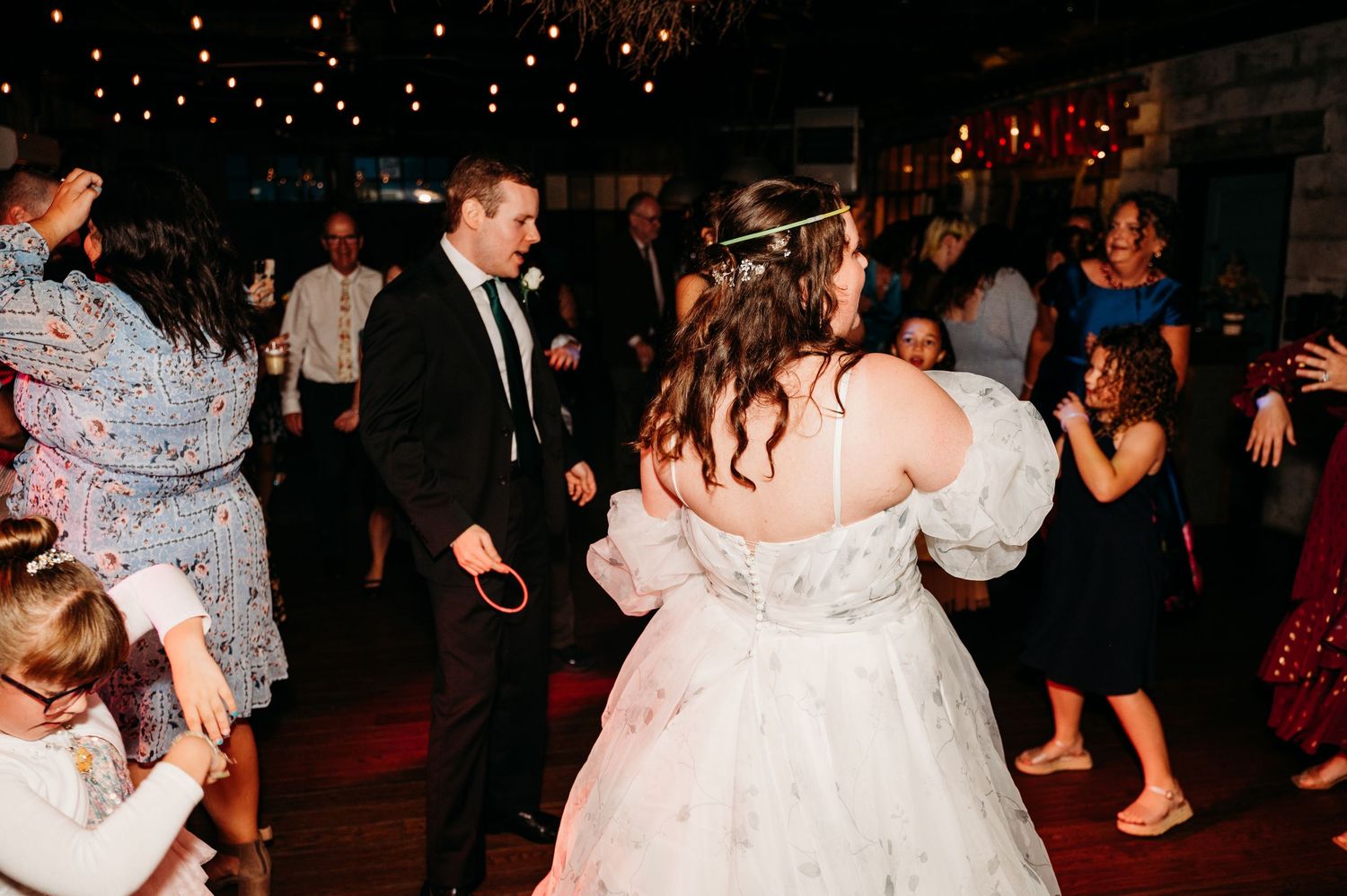 Wedding guests dance together at a dimly lit indoor reception with twinkle lights overhead.