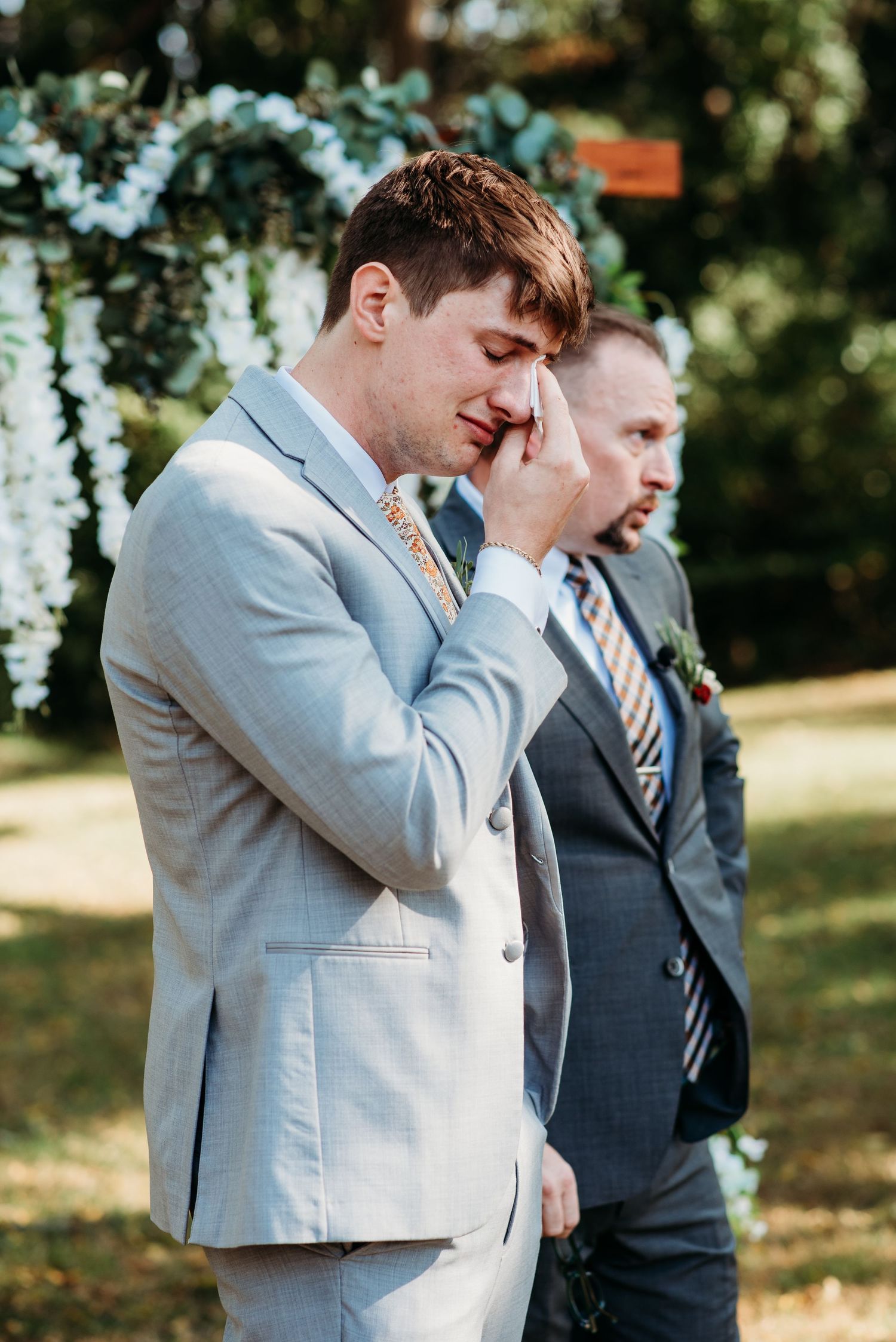 An emotional reaction during an outdoor wedding ceremony under a floral arch on a sunny day.