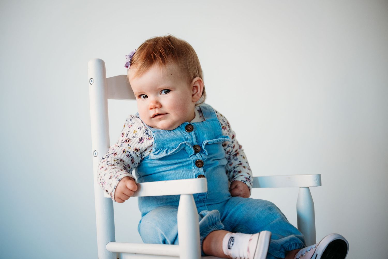 A baby in a bright blue sweater sitting on a white chair against a light blue wall.