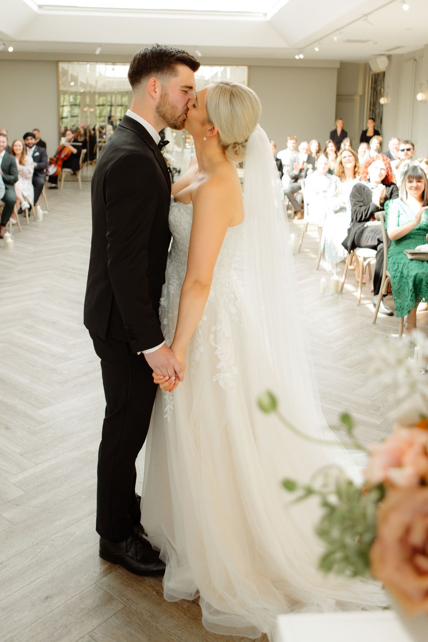 A tender kiss between newlyweds at their indoor wedding ceremony with guests seated in the background.
