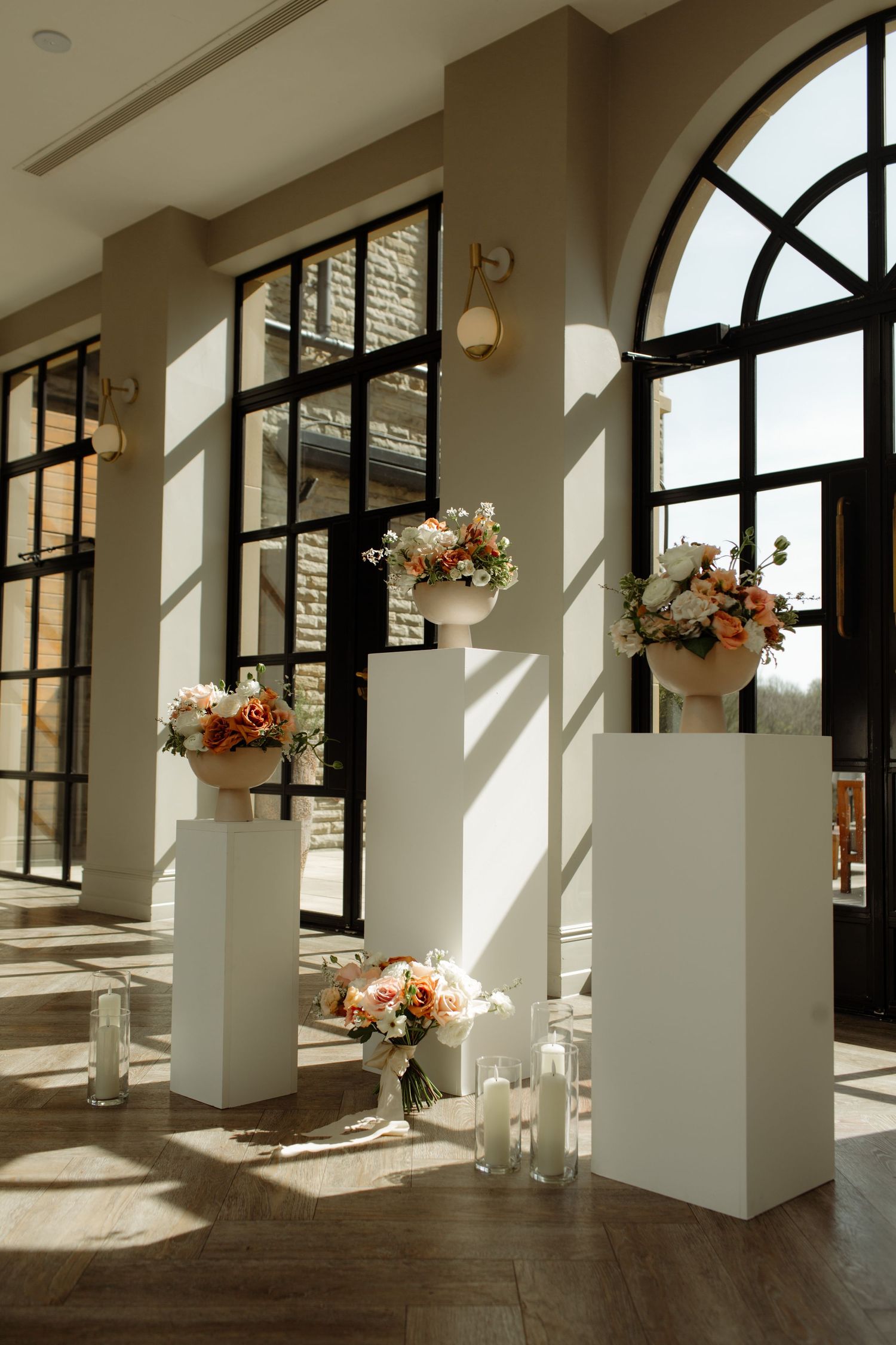 Elegant white floral arrangements on pedestals line a bright interior space with arched windows and hardwood floors.