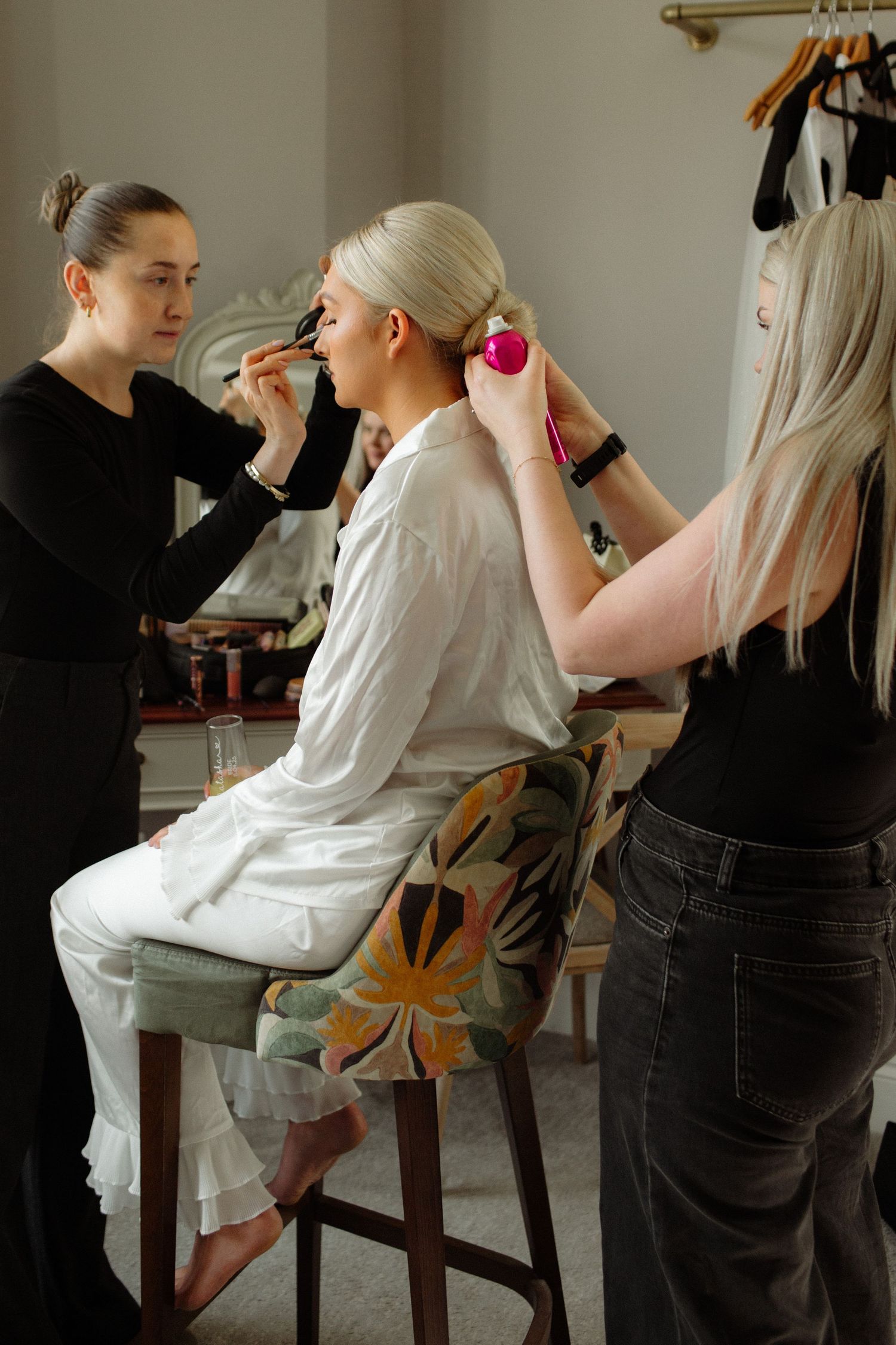 Two stylists work on a client's hair and makeup in a bright studio setting with clothes rack in background.