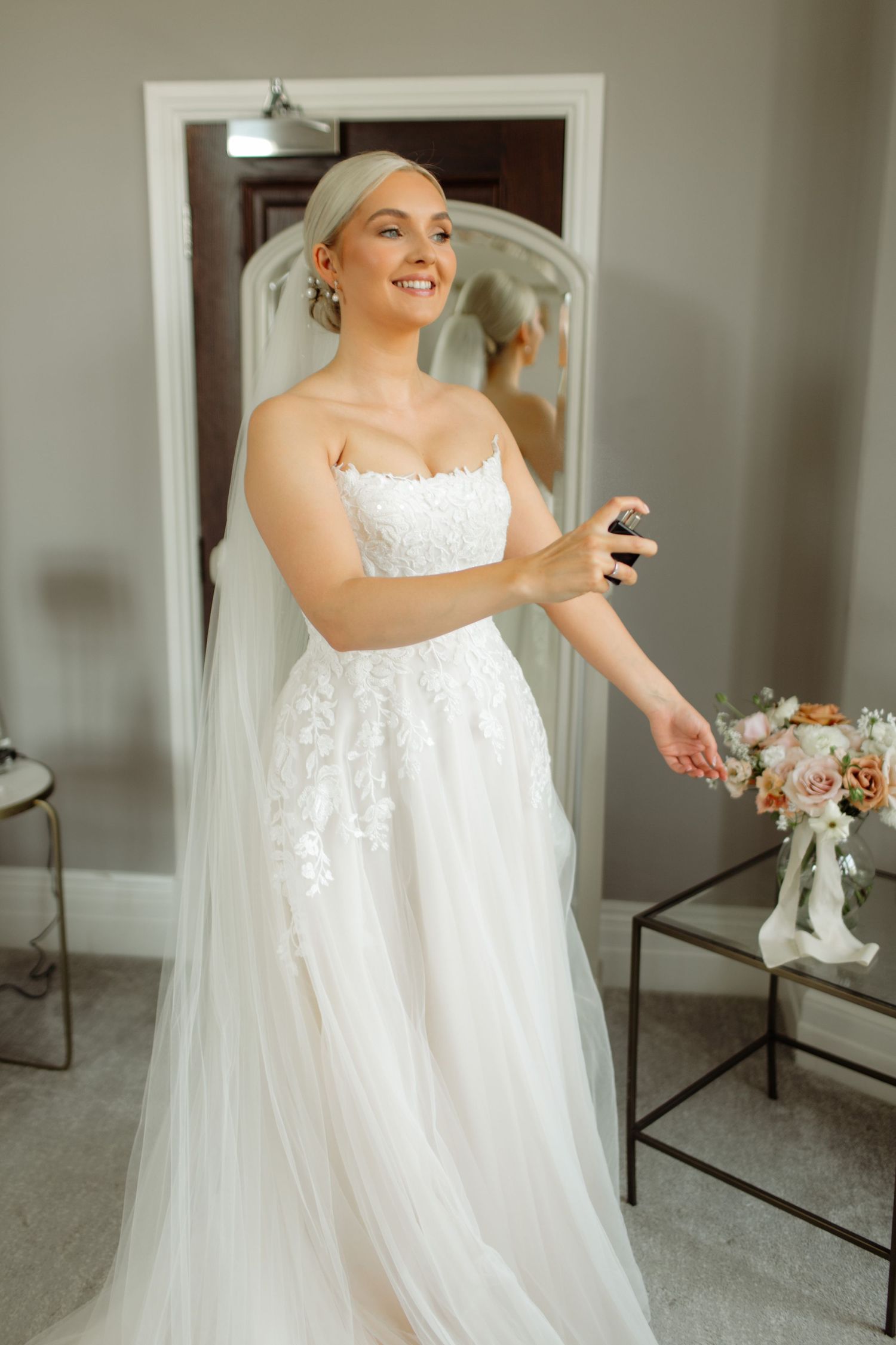 A bride in a white lace wedding gown and veil smiles while getting ready in a room with soft lighting.