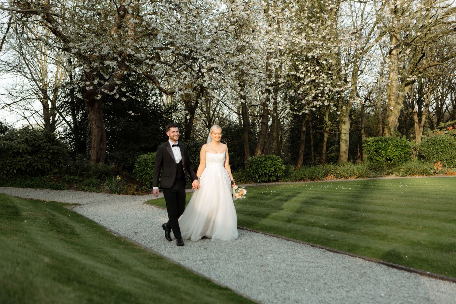 A couple walks along a gravel path through a lush garden with spring trees and manicured lawn.