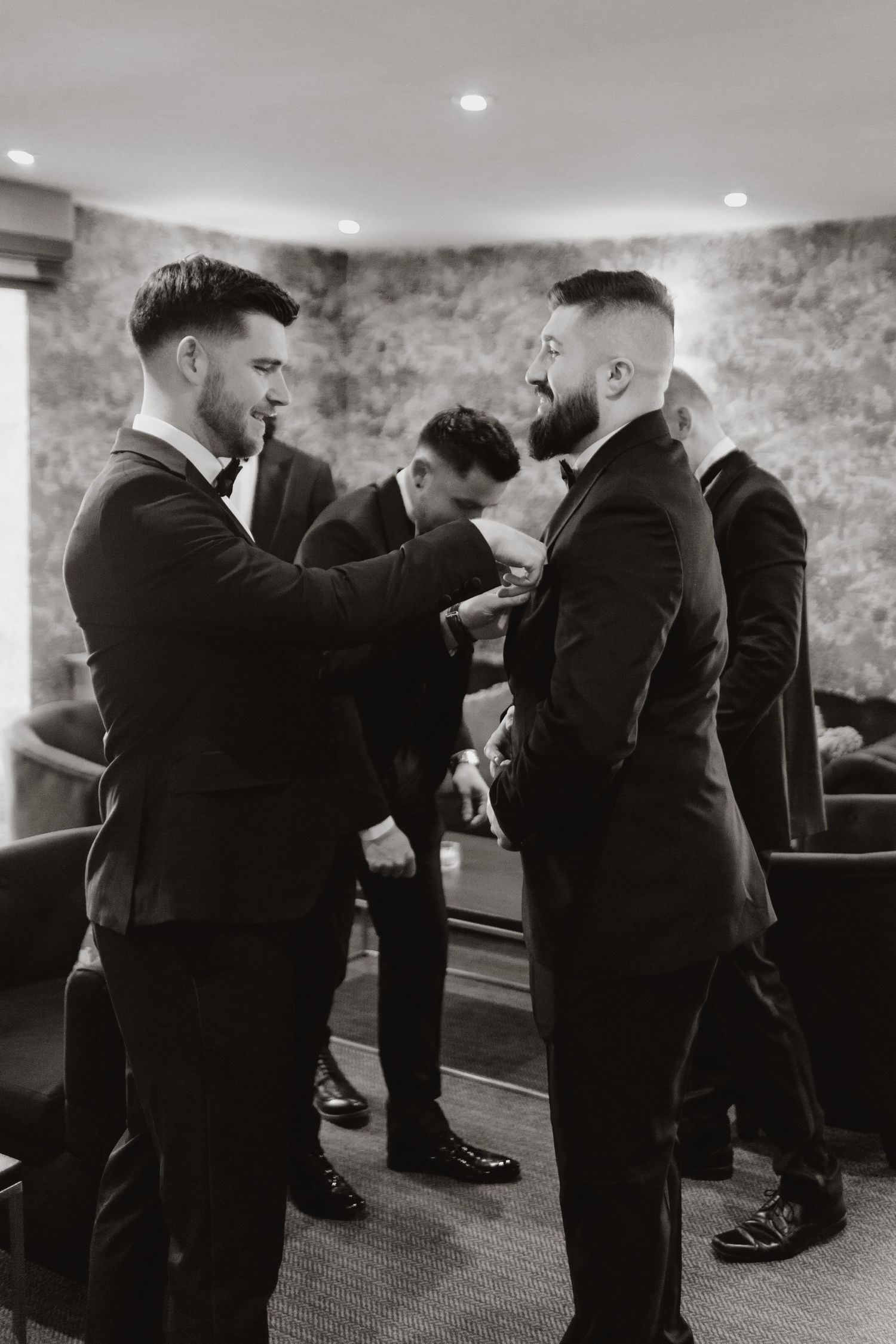 A black and white photo of groomsmen helping each other get ready before a wedding ceremony in a stone-walled room.