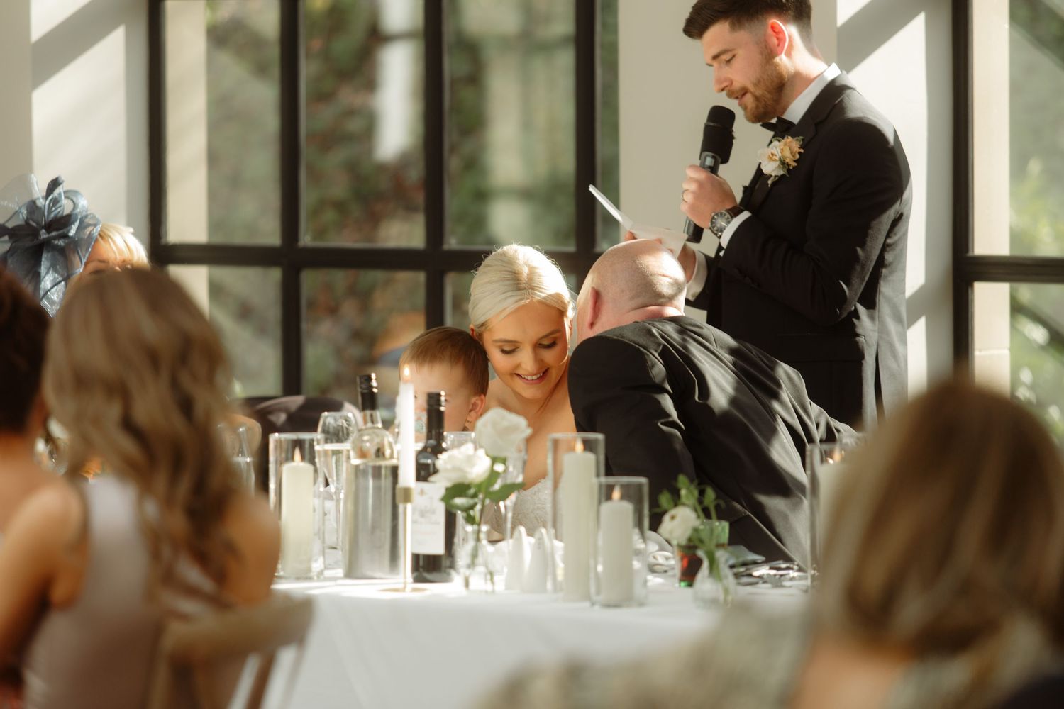 A speaker delivers a heartfelt speech at an elegant wedding reception with white table settings and candles.
