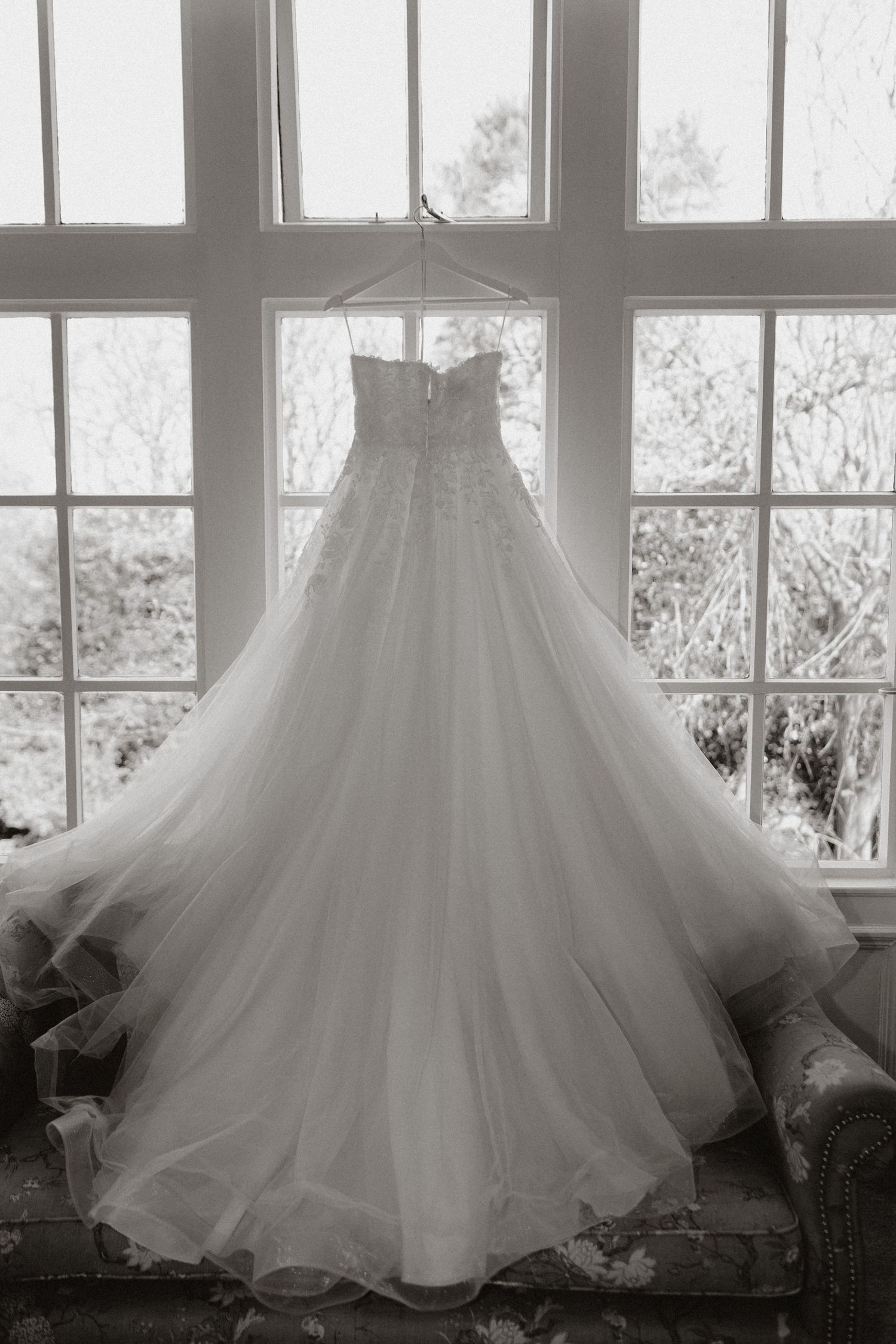 A row of elegant white wedding dresses with flowing tulle skirts silhouetted against bright windows in black and white.