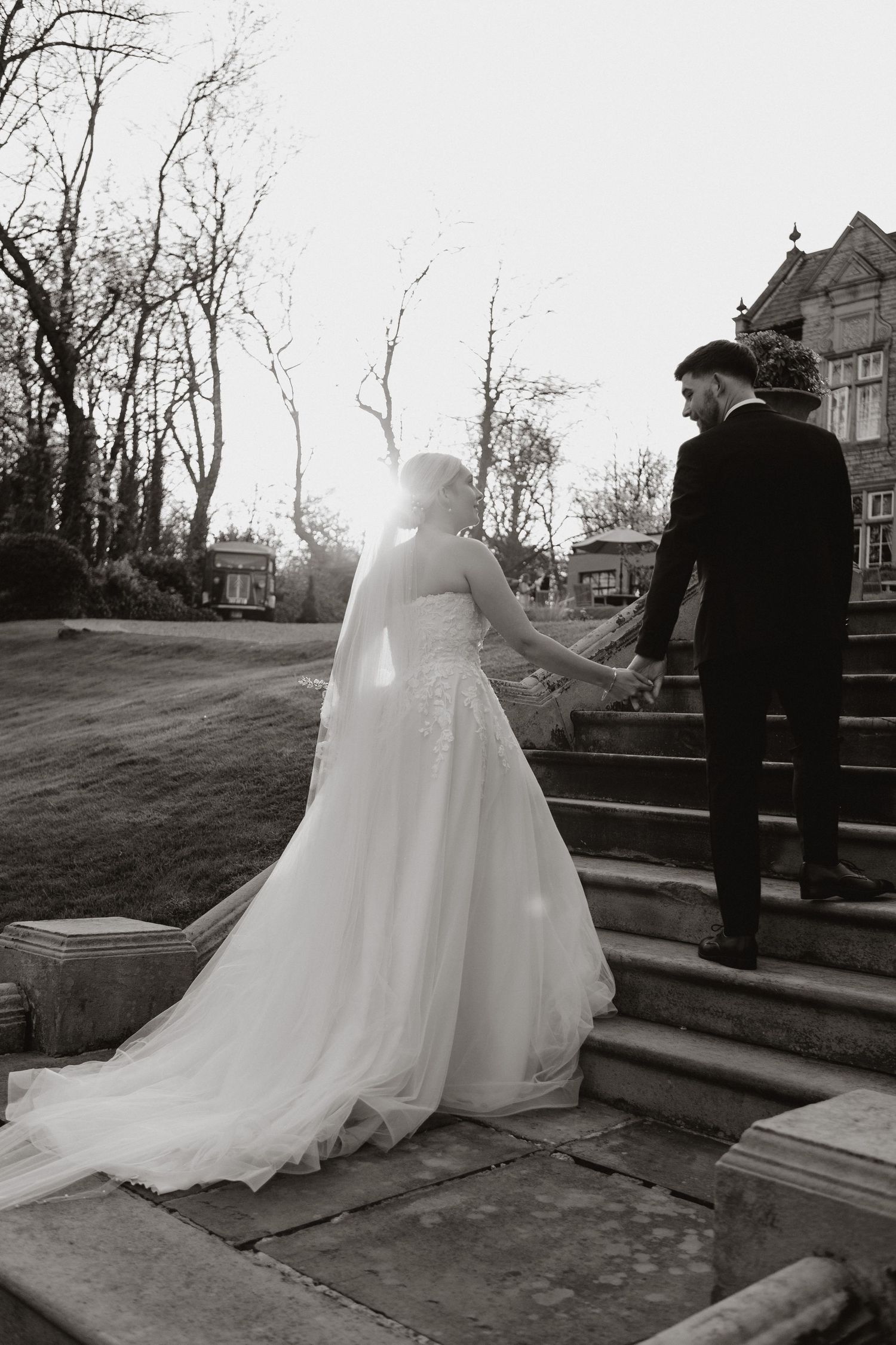 Black and white wedding photo showing a couple ascending stone steps together on their special day as sunlight peeks through trees.