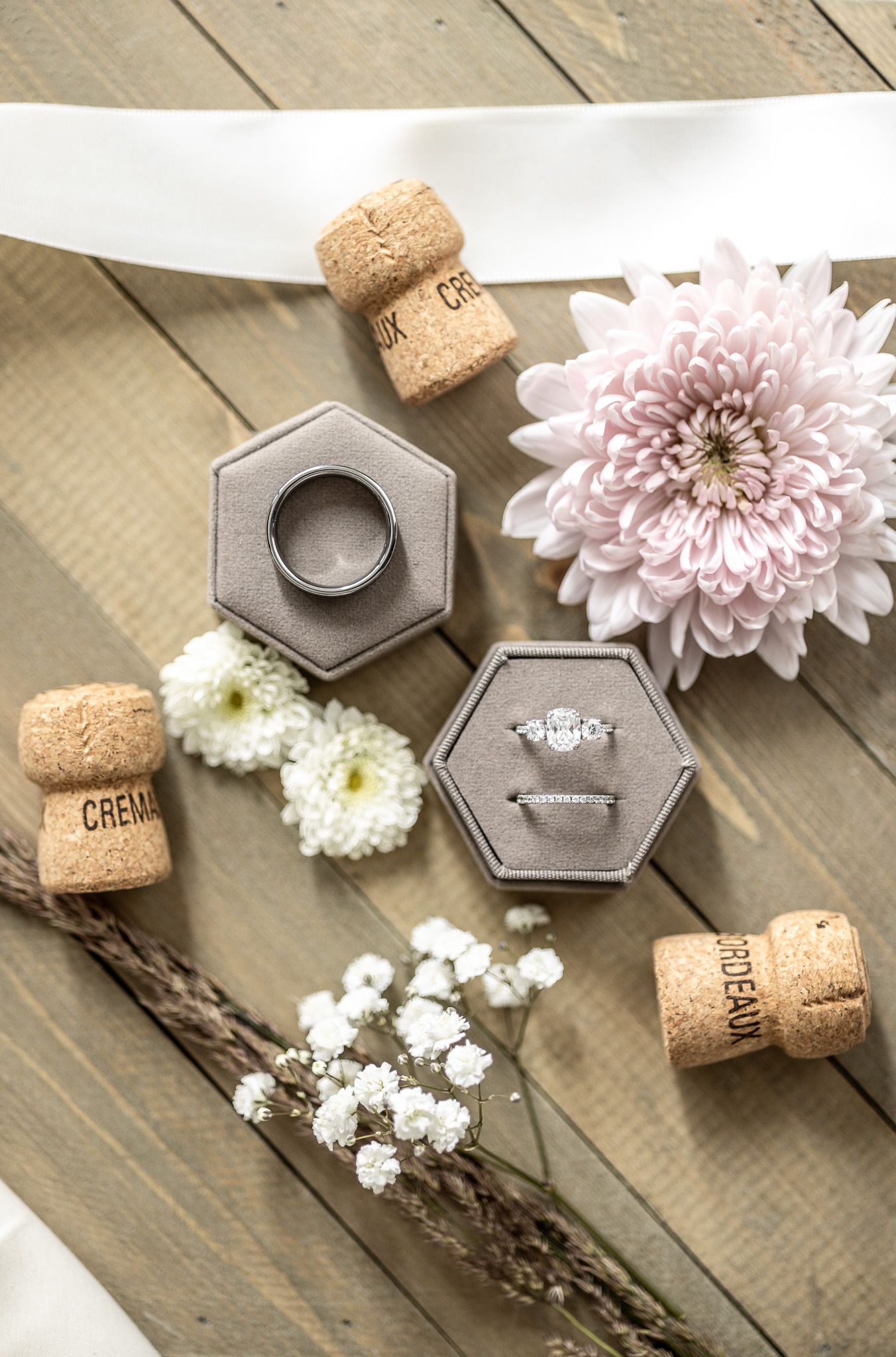 Wedding ring and engagement ring displayed in hexagonal jewelry boxes with wine corks and soft pink flowers on wooden surface.