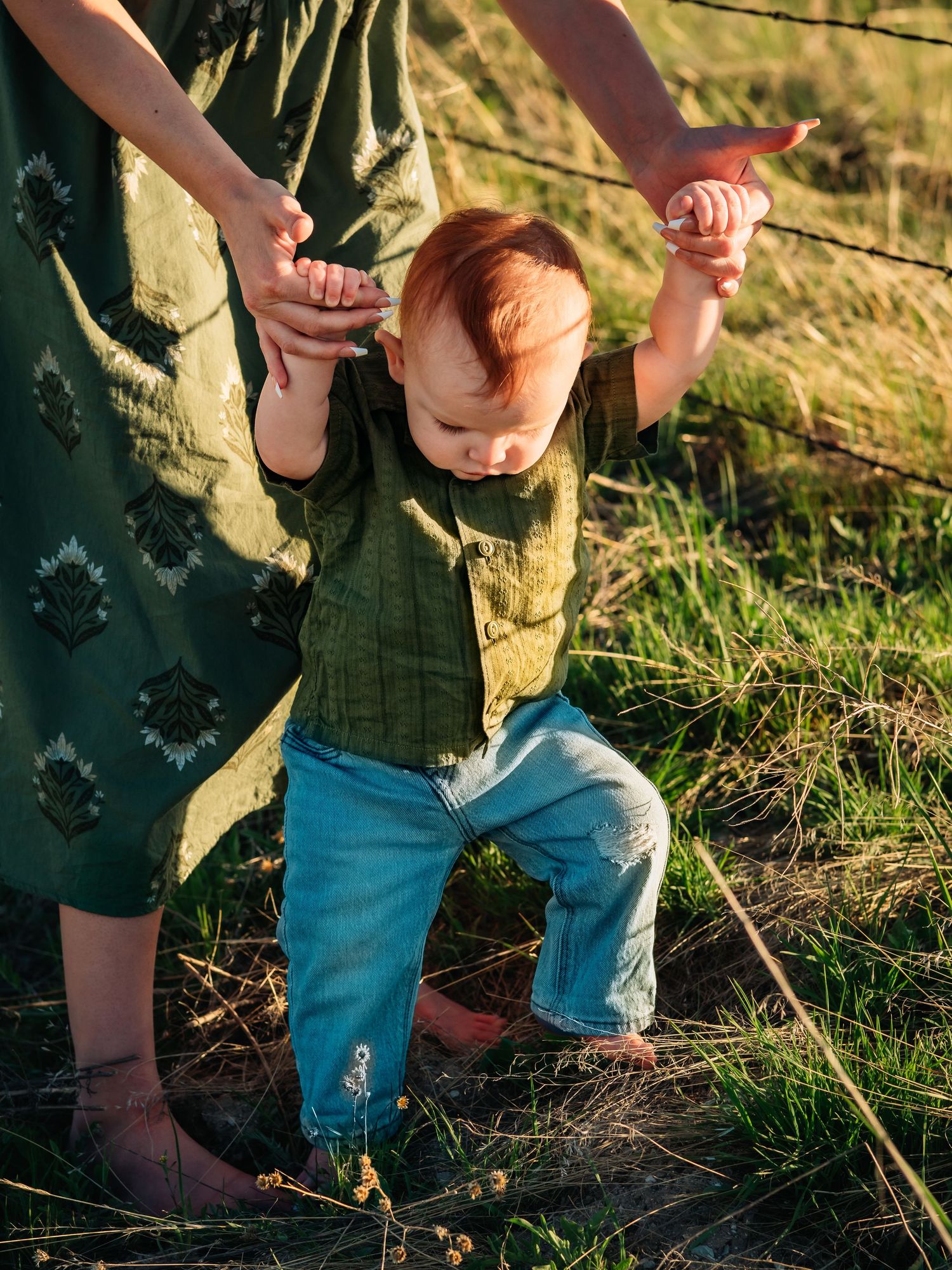 Small child in green shirt and jeans learns to walk while holding hands in sunny field.