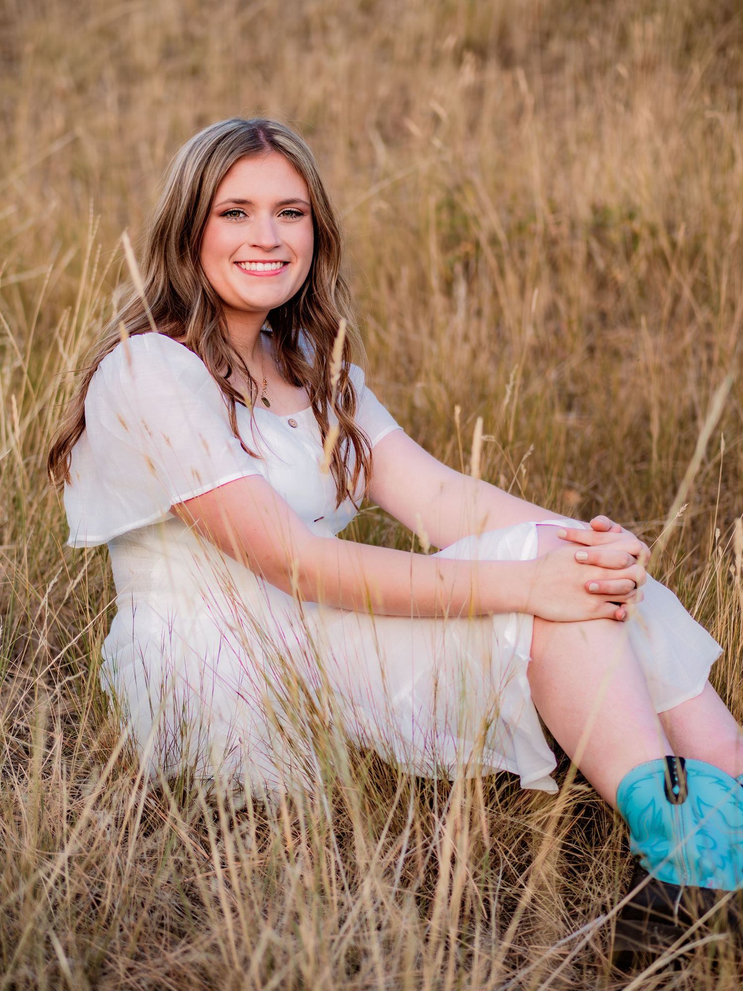 Person in white dress and turquoise boots sits in golden grass field during sunset.