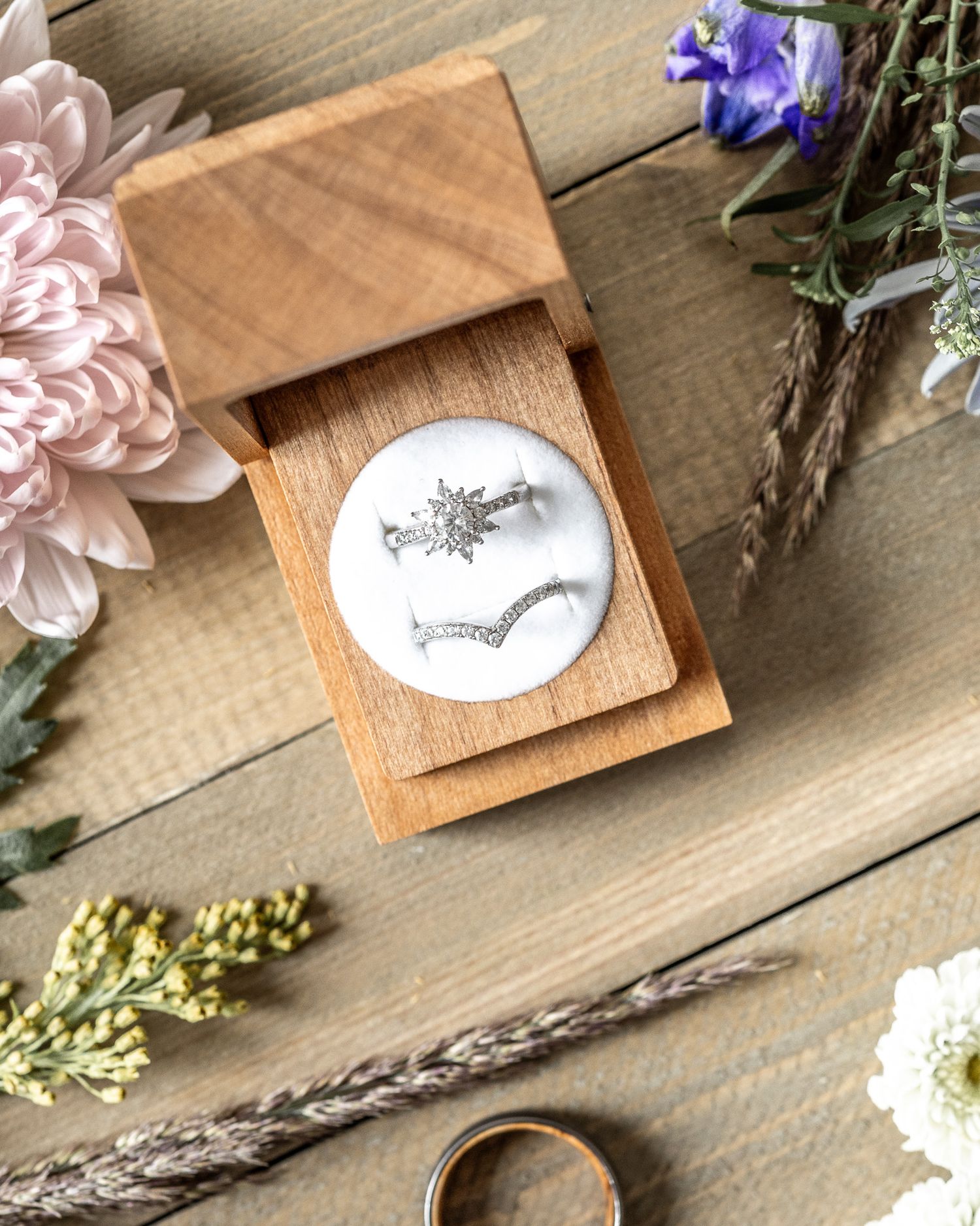 Wooden ring box with floral illustration displayed among pink flowers, purple iris, and dried wheat stems on wooden surface.