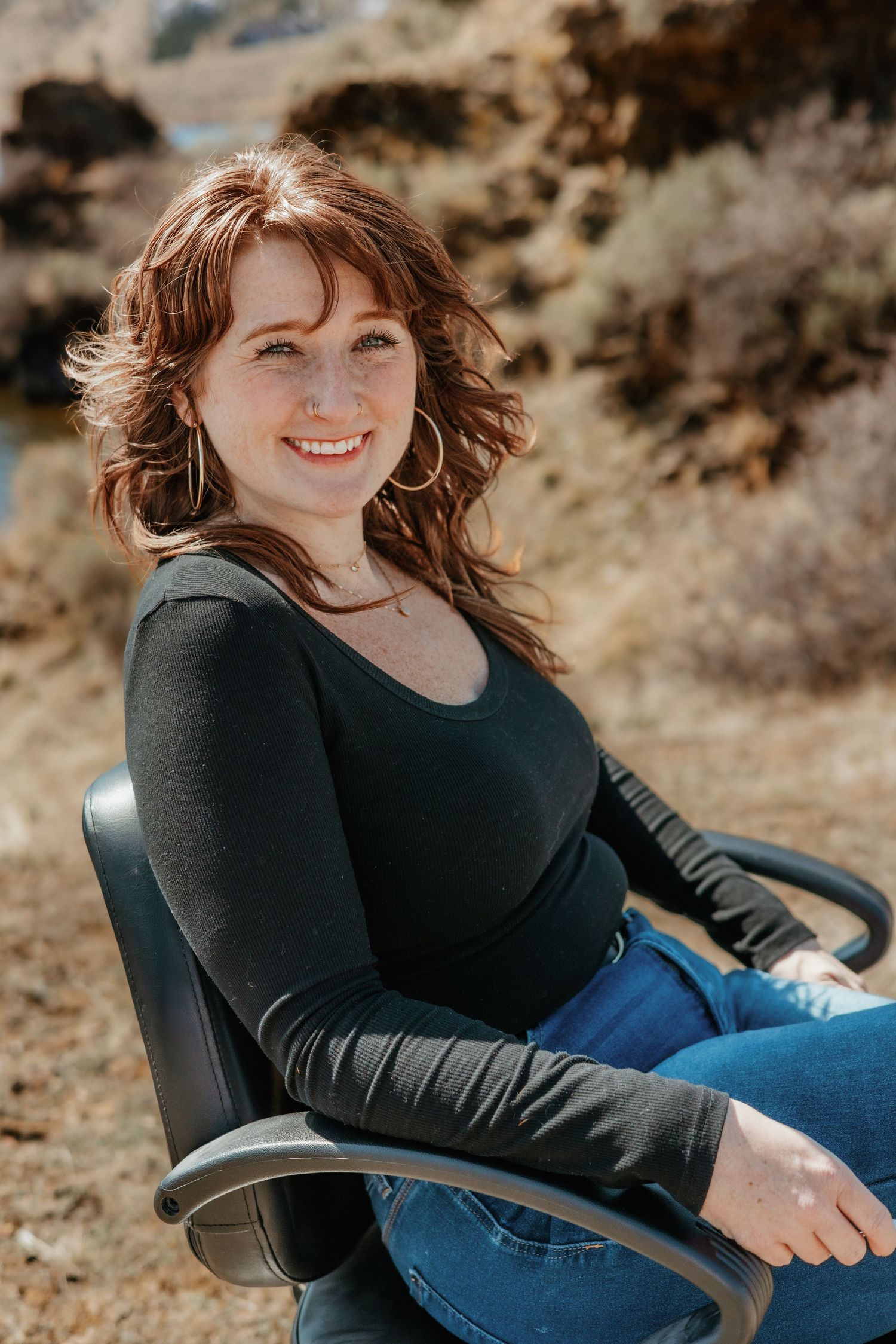 Person wearing a black long-sleeve top and jeans sits in a chair against a natural outdoor backdrop.