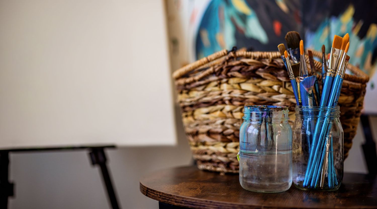 Art supplies arranged with paintbrushes in mason jar and woven basket on wooden surface.