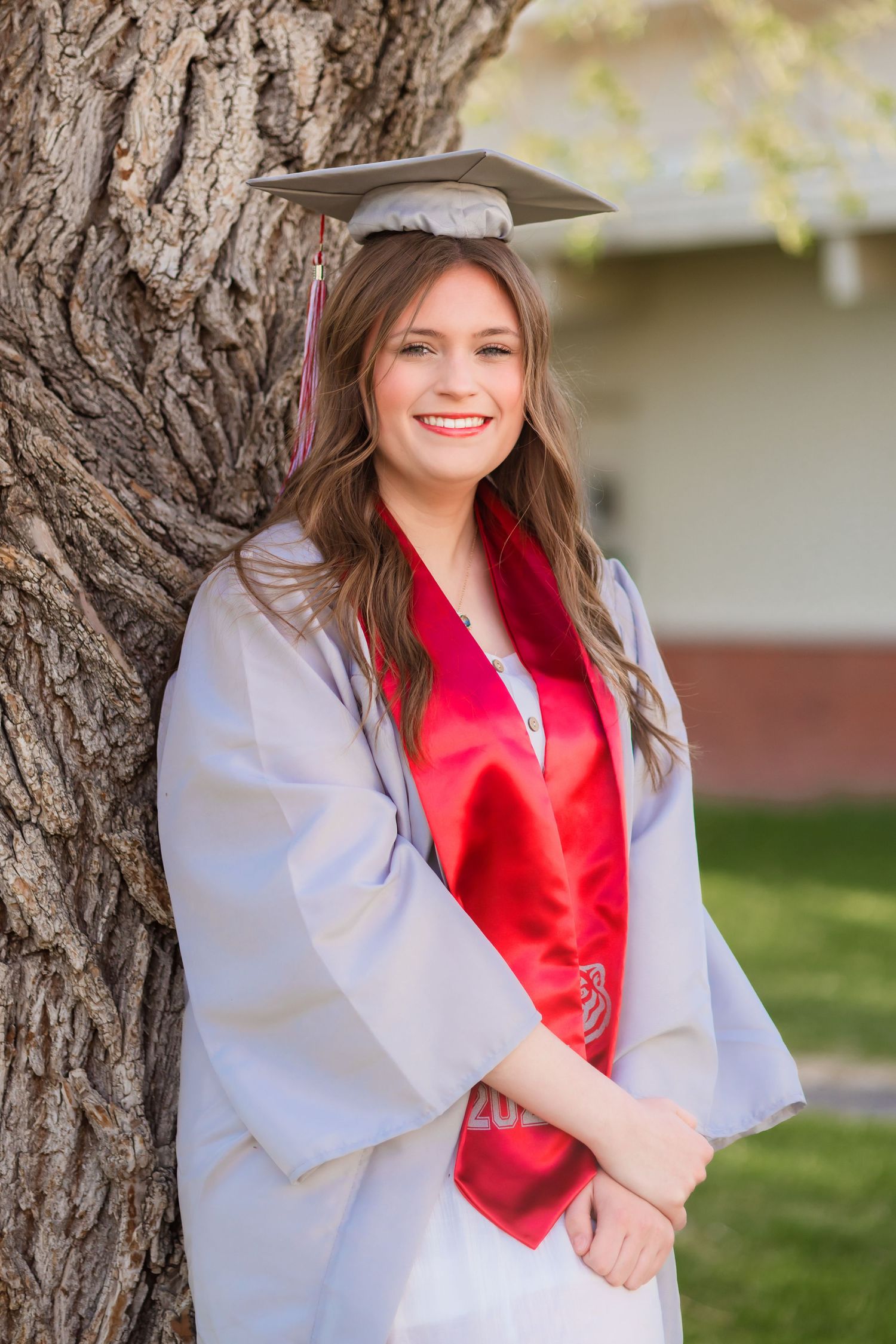 A graduate in cap and gown smiling by a tree trunk.