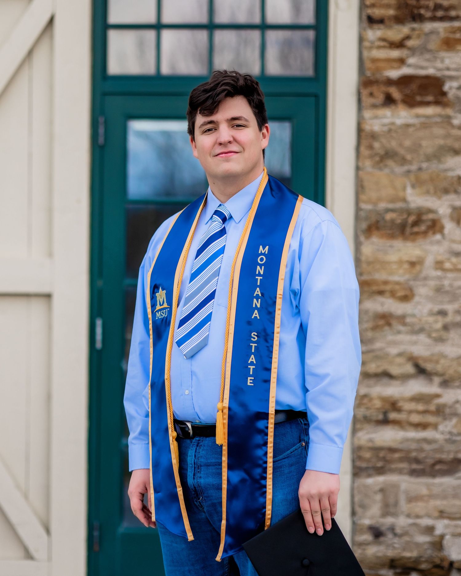 Graduate in blue dress shirt and striped tie wearing honor cords against stone wall.