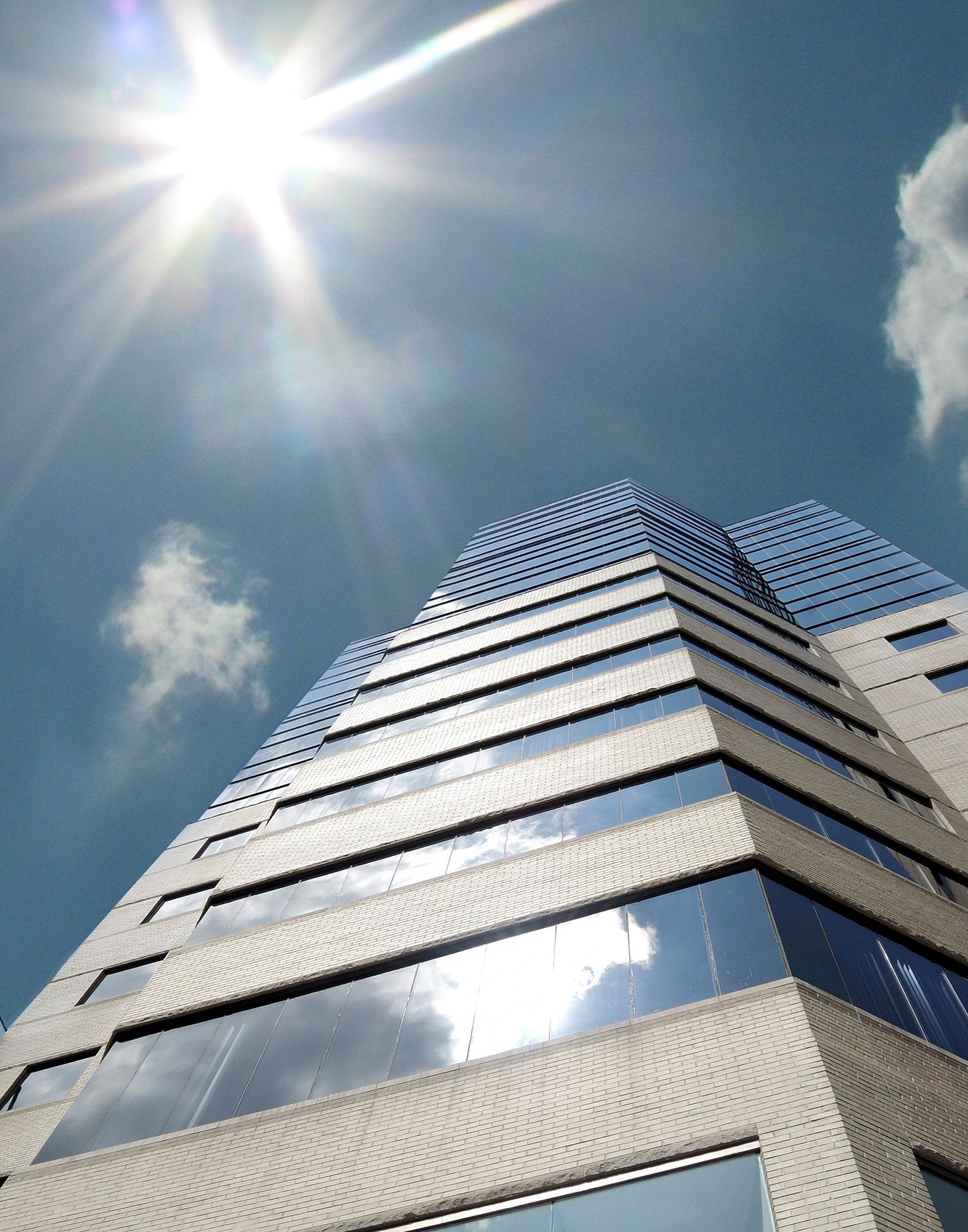 Modern office building with reflective glass windows photographed from low angle against bright sunlight and blue sky with clouds.