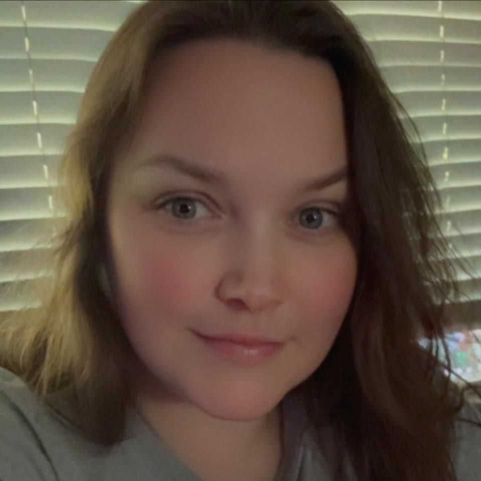A selfie portrait with brown hair and natural makeup against a background of venetian blinds.