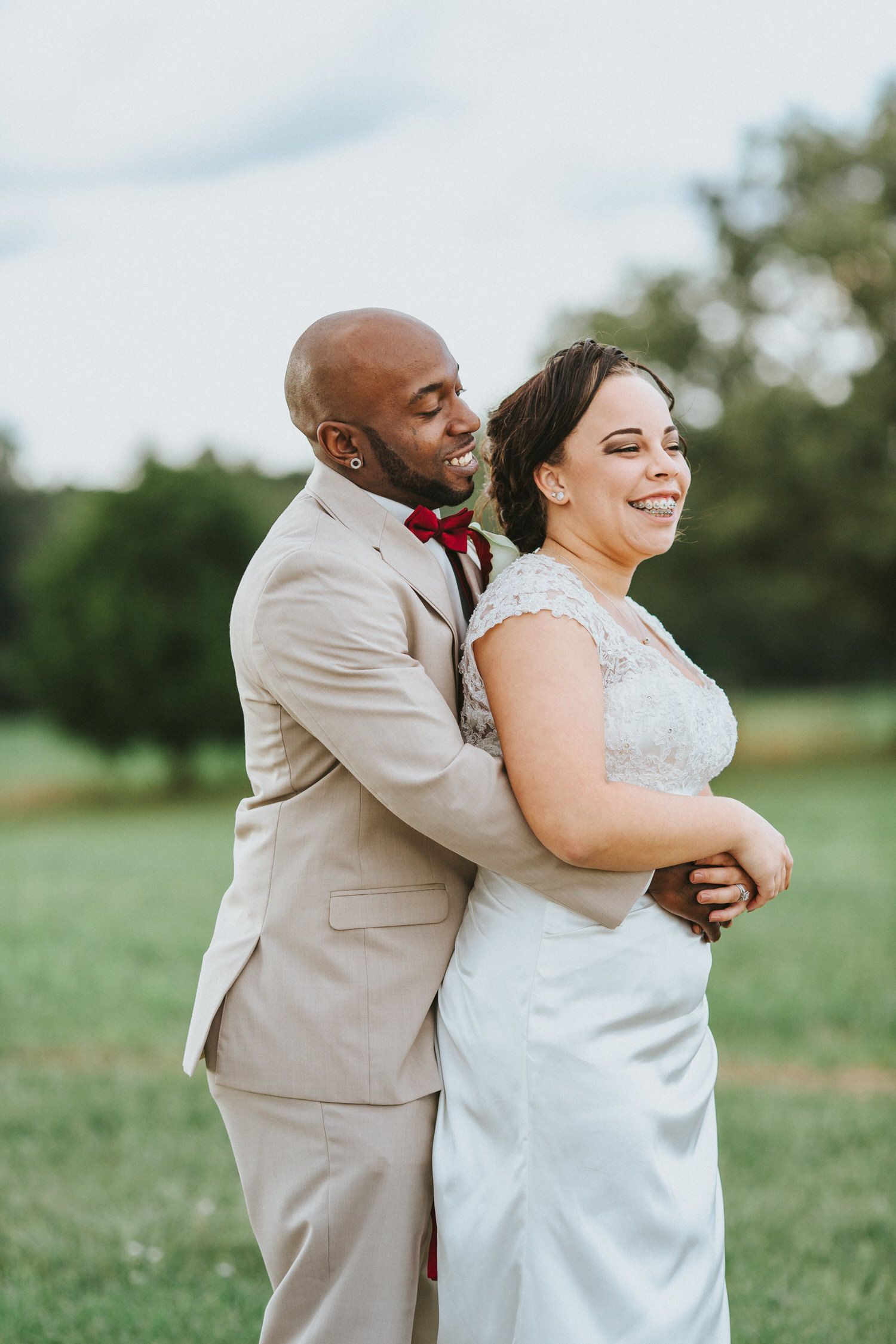 A happy couple embraces outdoors during their wedding celebration, wearing a beige suit and white lace gown.