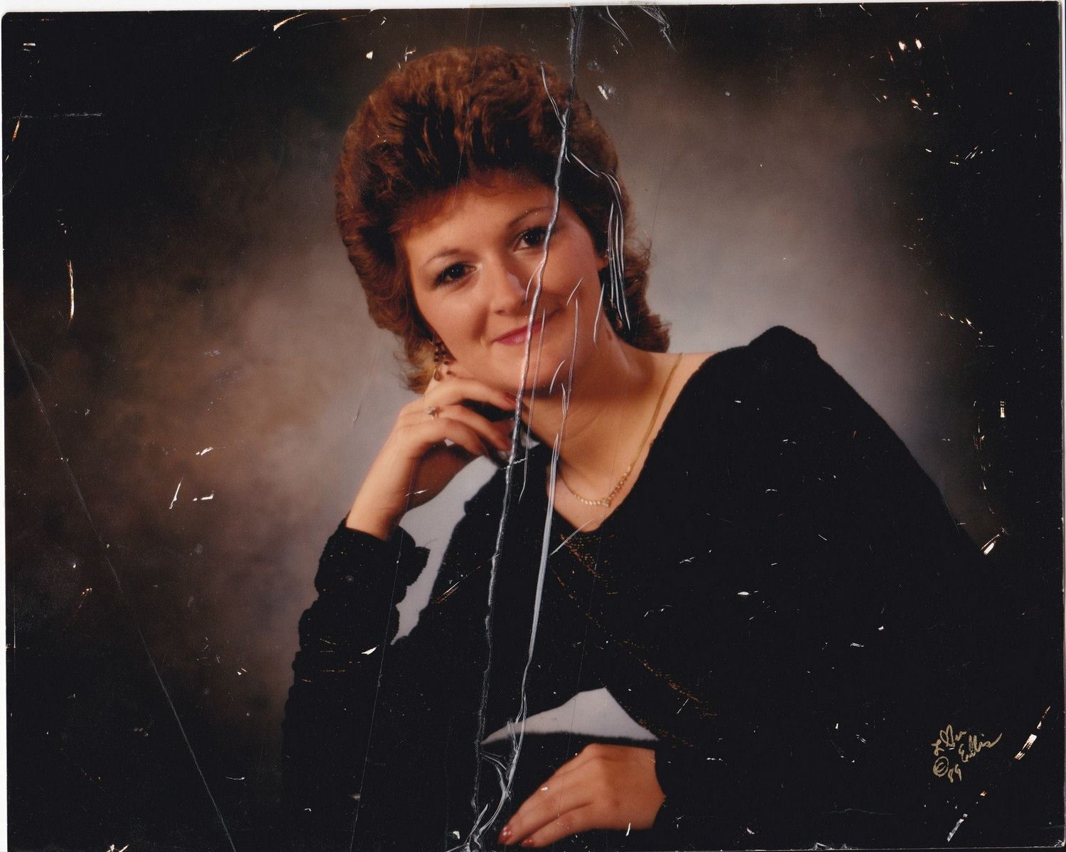Studio portrait photograph showing someone in a black outfit posing against a dark background with moody lighting.