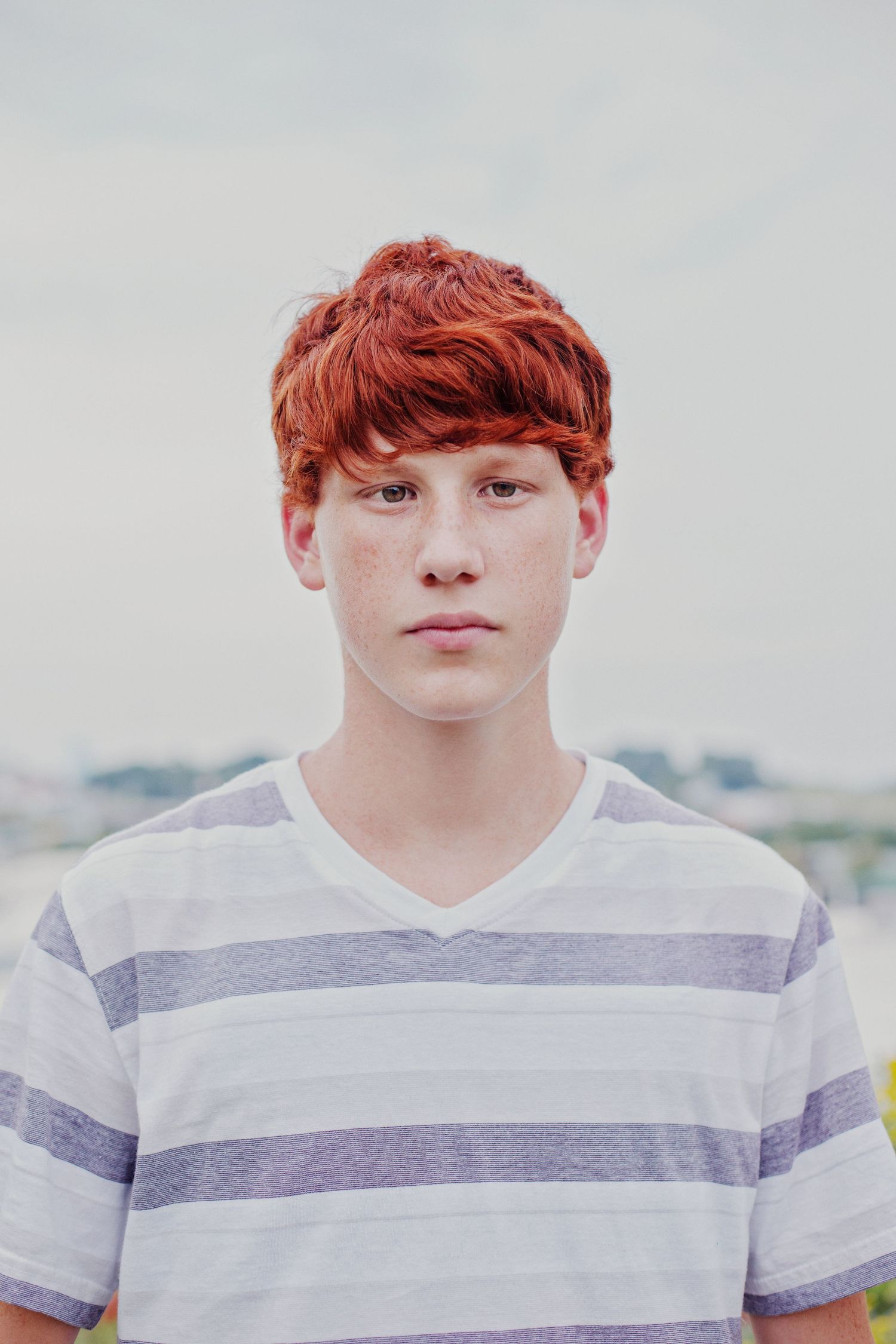 Portrait of a young person with red hair wearing a white and gray striped t-shirt against a bright background.