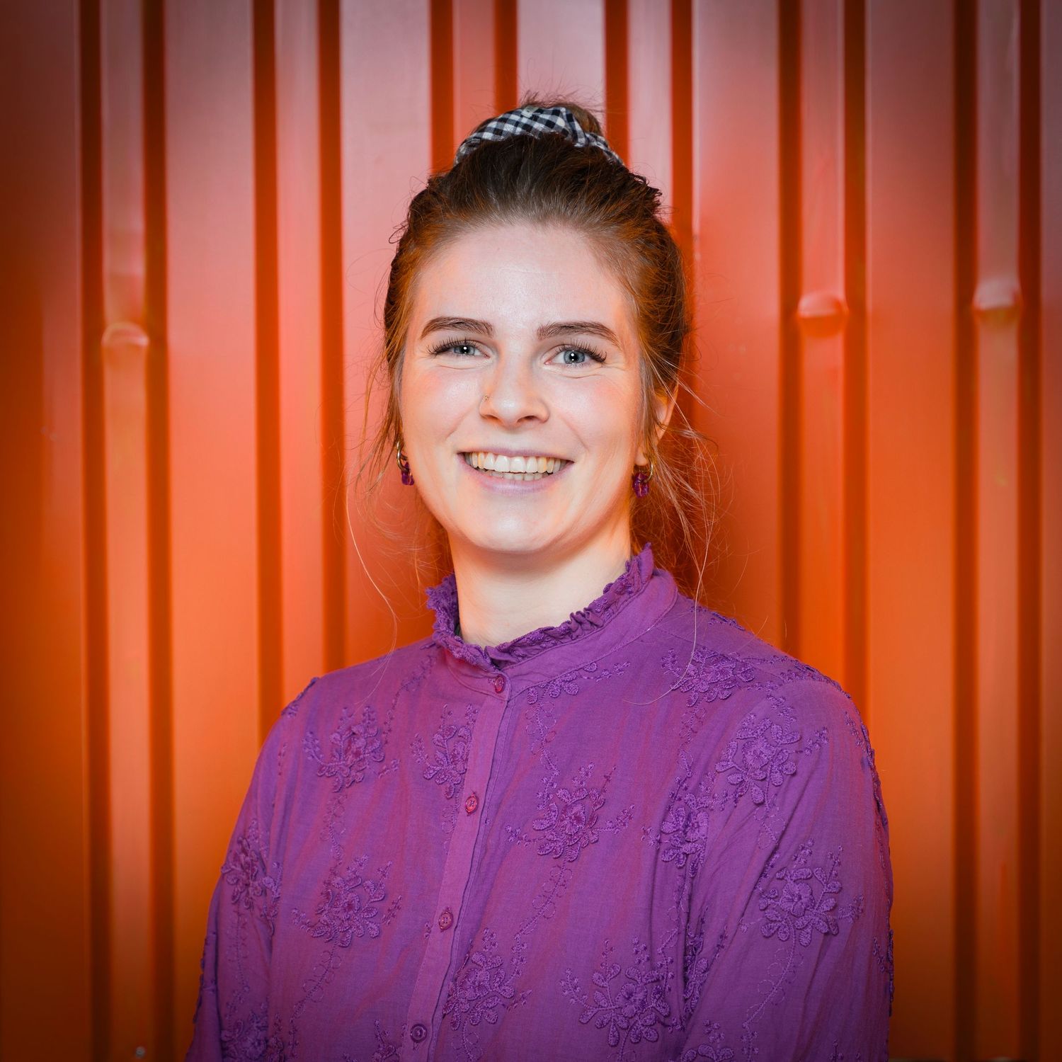 A smiling person in a purple collared top stands against an orange corrugated metal wall background.