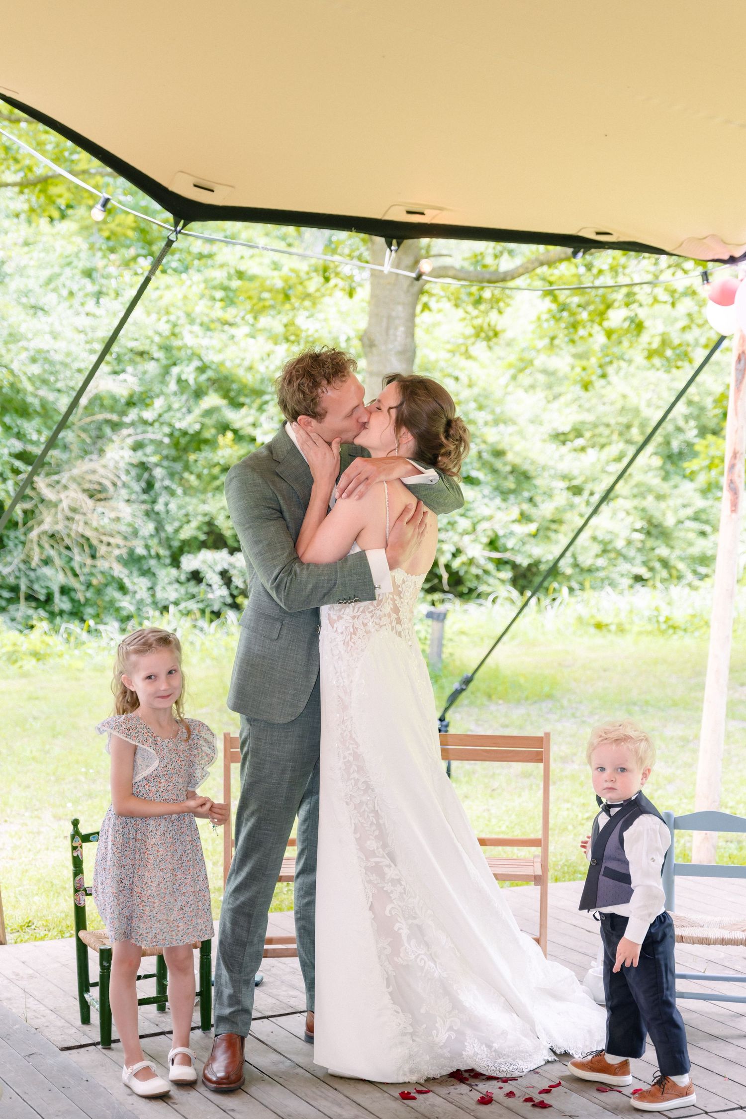 A romantic wedding moment shared under a canopy with young attendants looking on at the outdoor ceremony.
