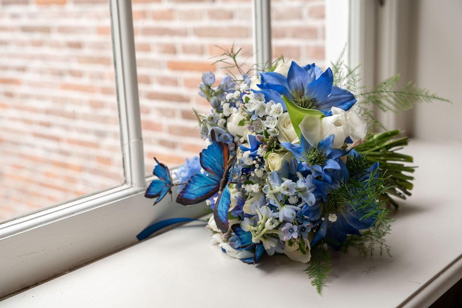 Bridal bouquet with vibrant blue delphiniums and white flowers rests on a windowsill with brick wall backdrop.