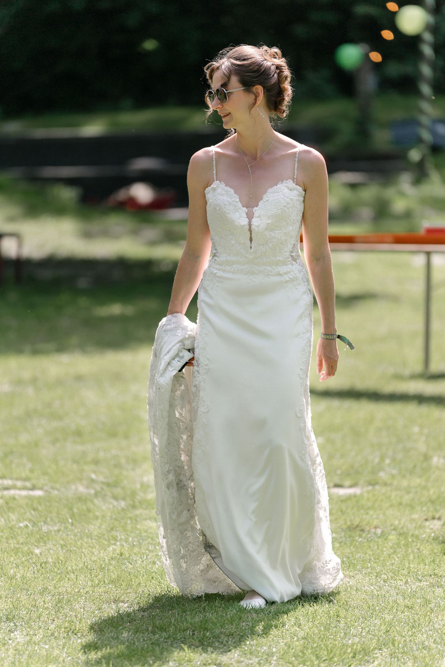 Beautiful bride in a sleek white spaghetti strap wedding gown with lace details walks gracefully across sunny garden lawn.