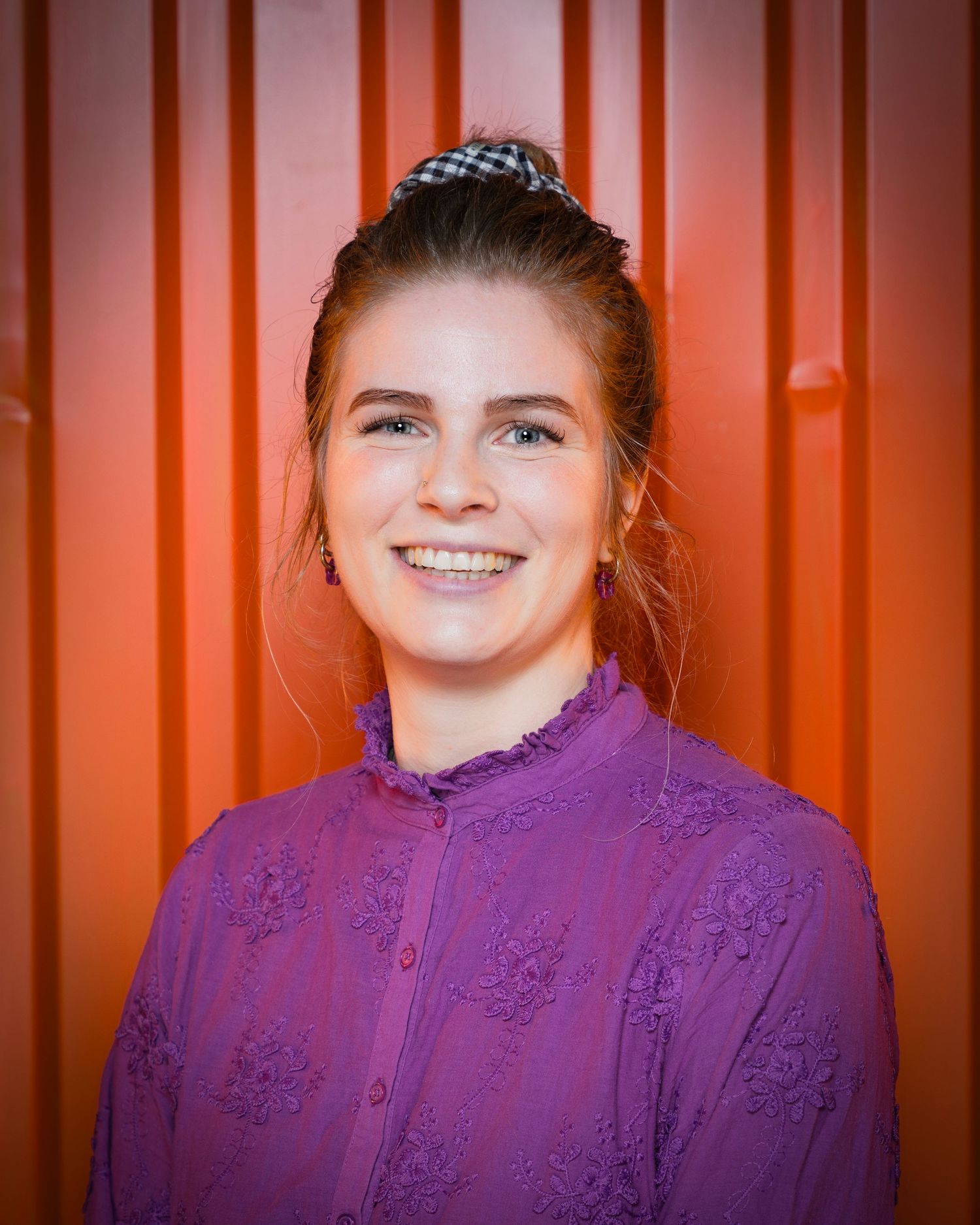 Portrait of person in purple embroidered blouse smiling warmly against an orange striped background.