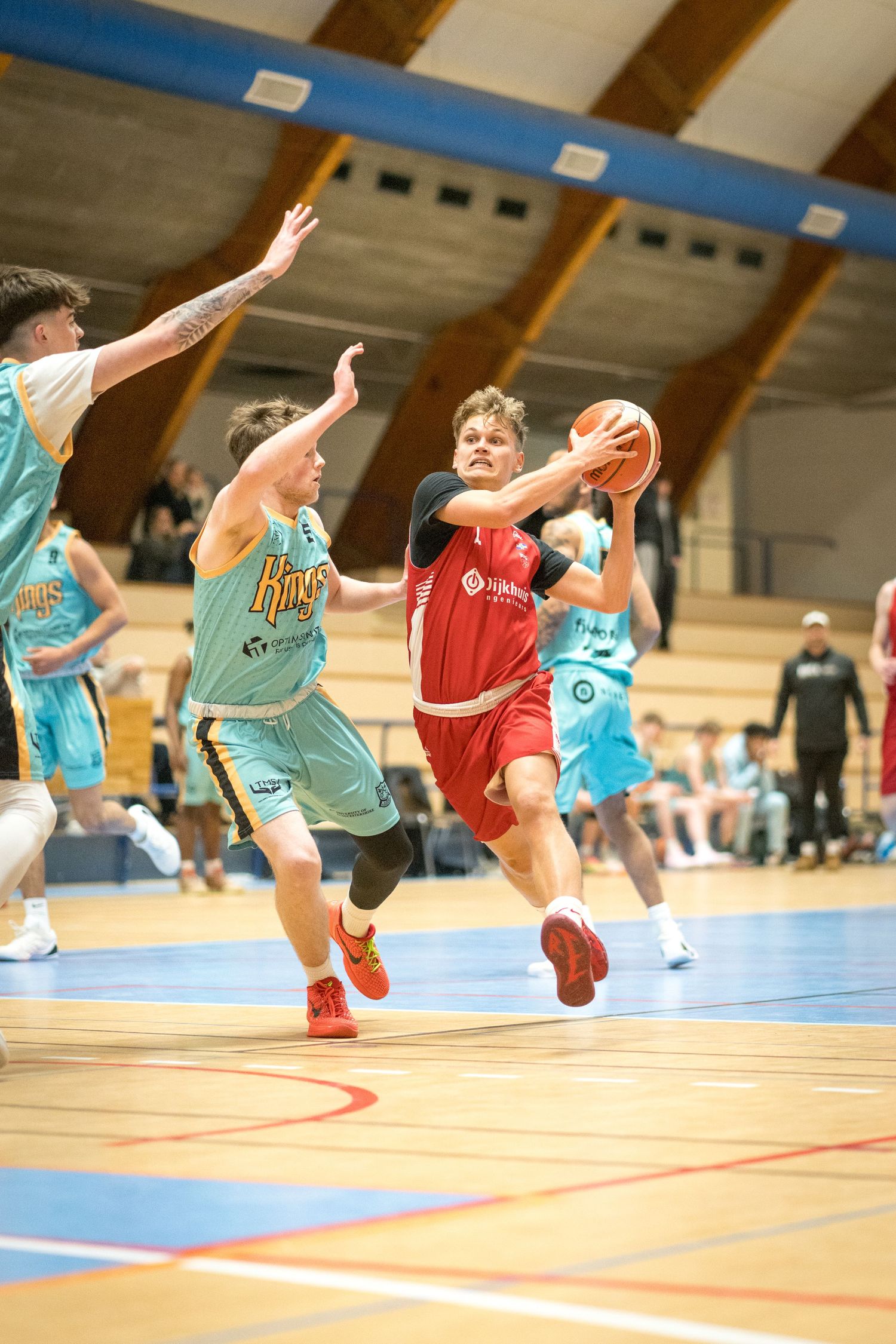Basketball players compete during an intense game with players in light blue and red uniforms on indoor court.