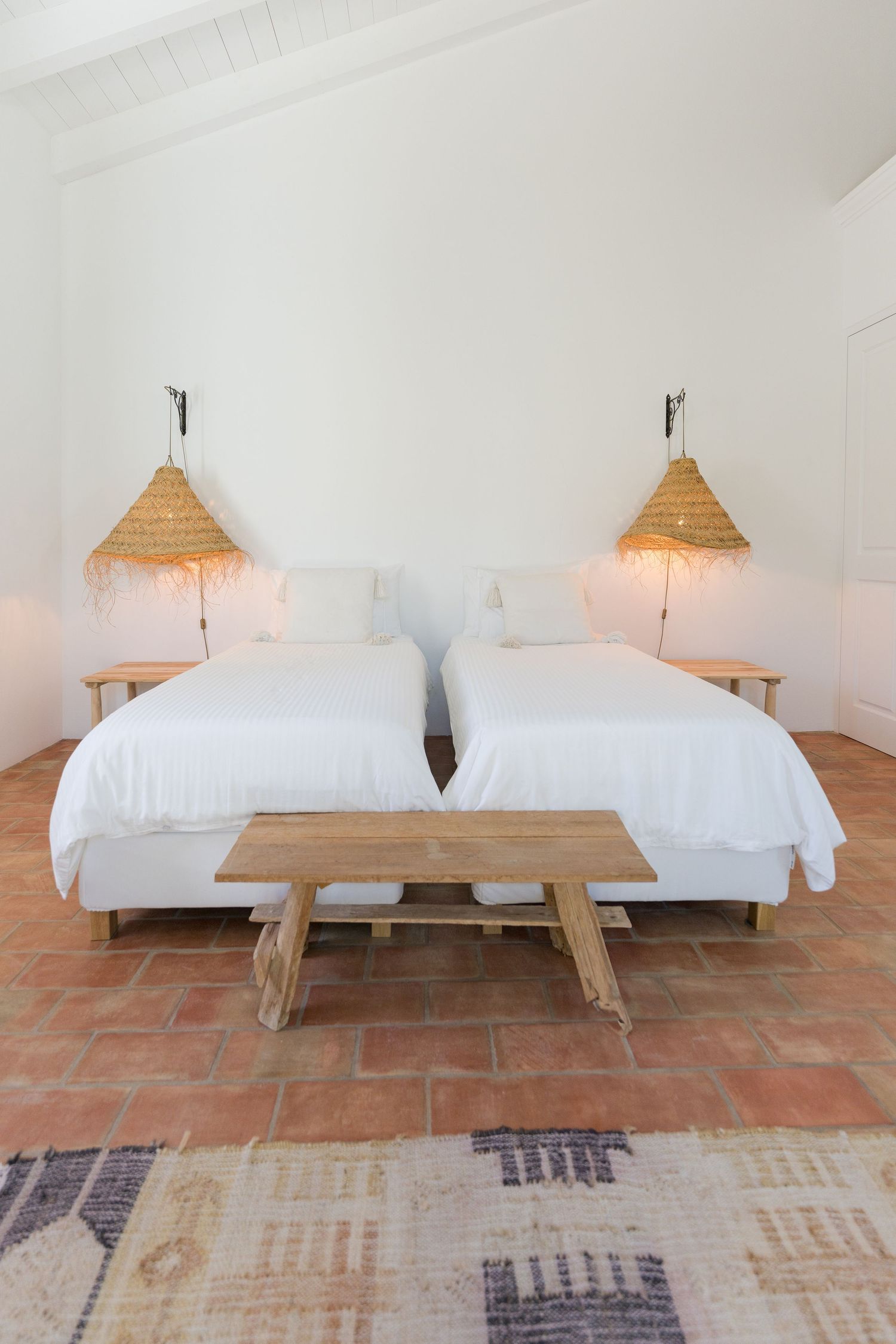 Twin bedroom with woven bamboo pendant lamps, white linens, and rustic wooden bench on terracotta floors.