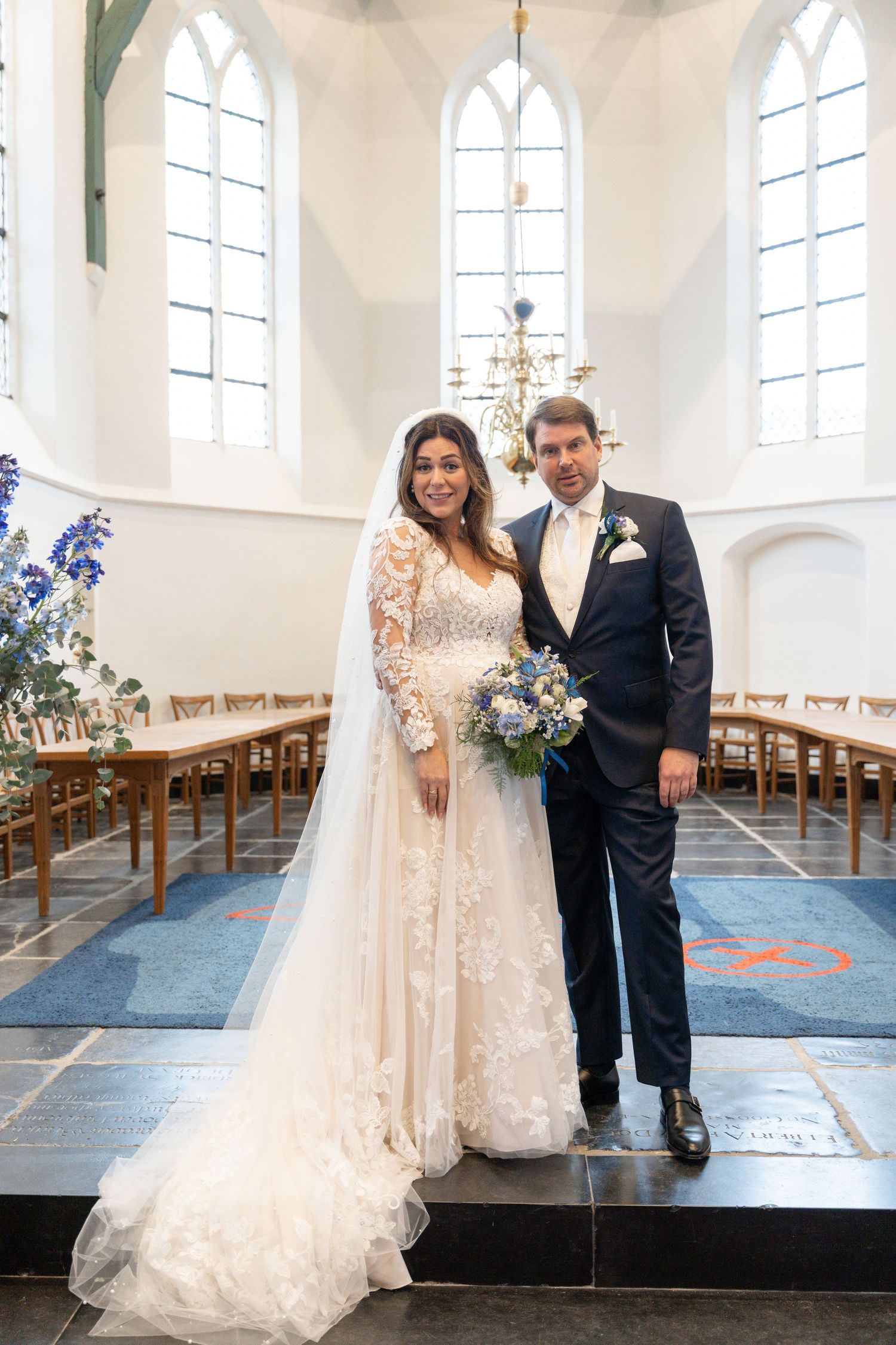 A couple in wedding attire stands together in a bright church with gothic windows and wooden pews.