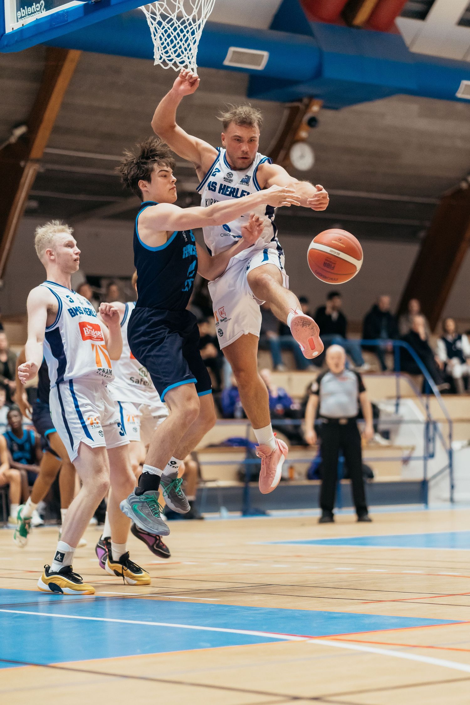 Basketball players collide mid-air during an intense game play on a bright blue court with spectators watching.