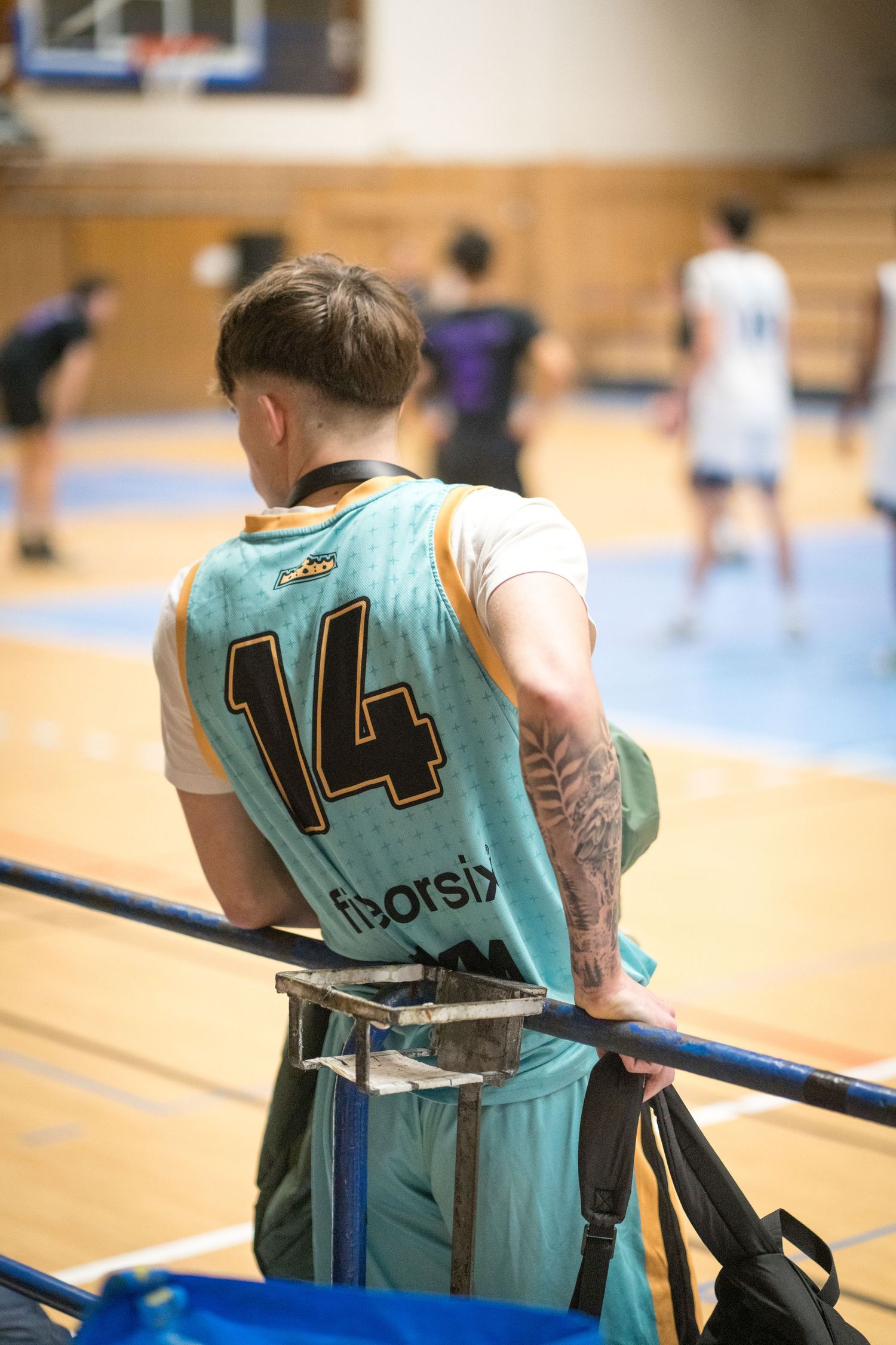 Basketball player wearing jersey number 14 in light blue uniform sits on sideline during indoor game.