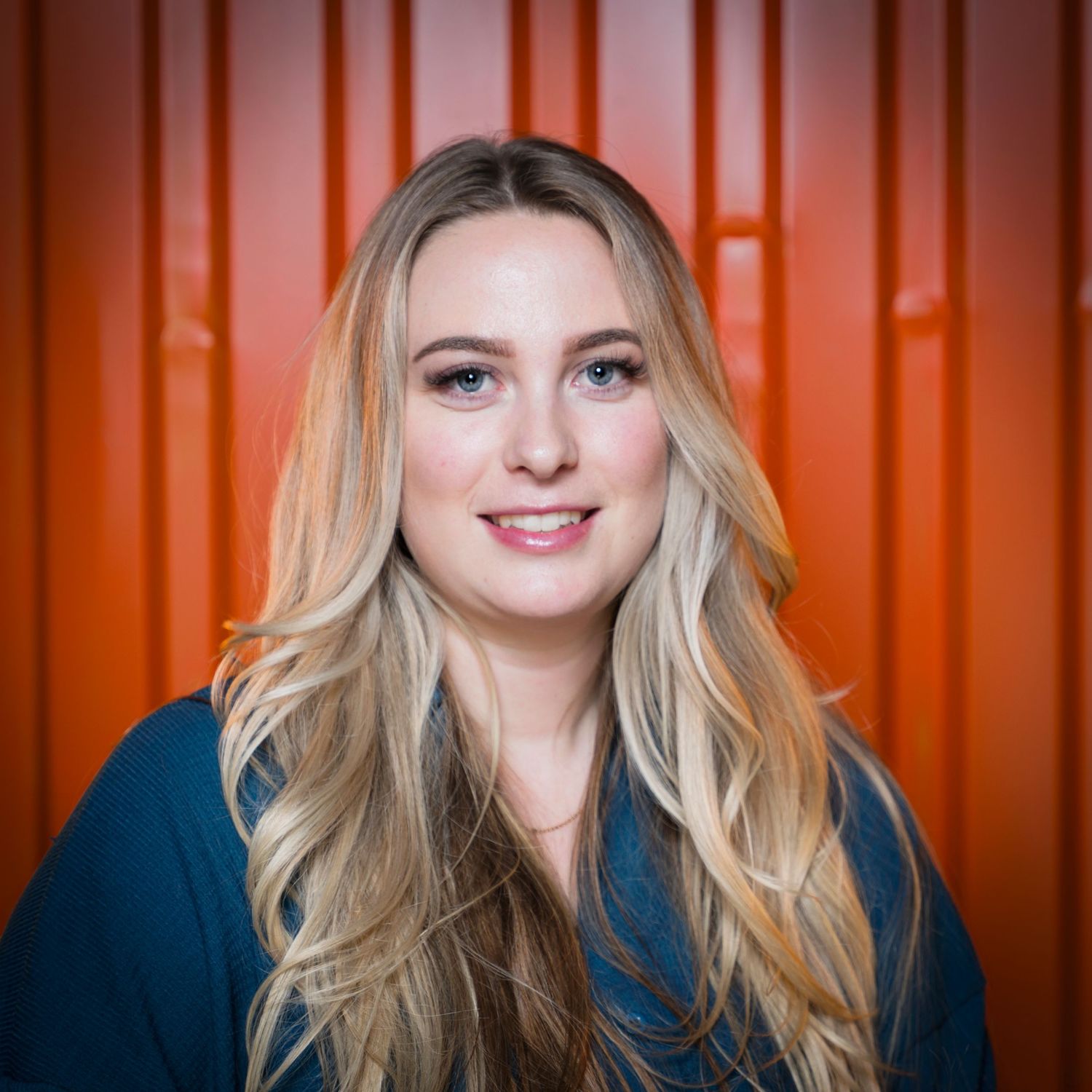 Professional headshot of a person with long blonde hair smiling warmly against an orange background while wearing a navy sweater.