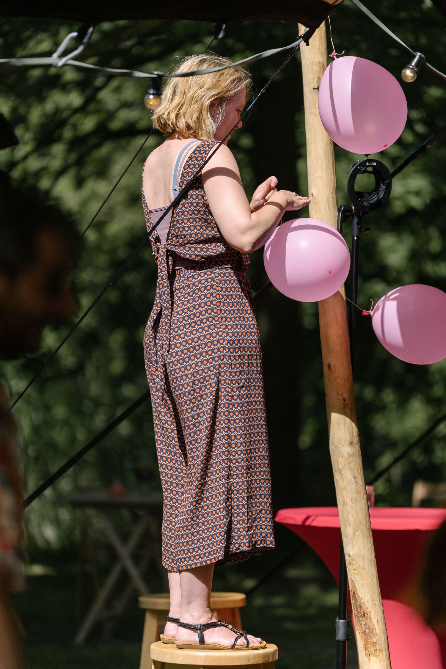 Person in patterned dress stands on stool hanging pink paper lanterns at outdoor summer party setup.