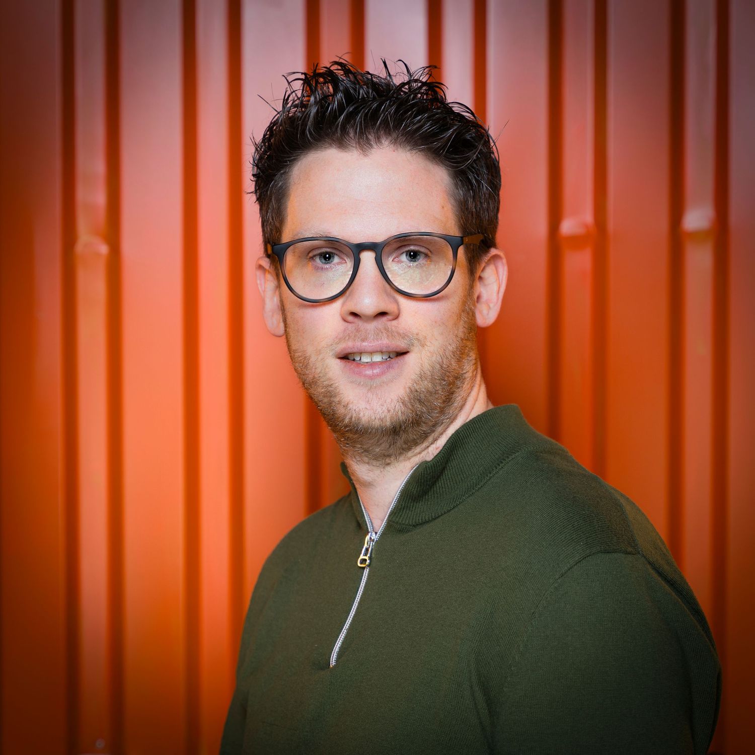 Portrait of a person with dark-rimmed glasses and stubble wearing an olive green zip-up top against an orange background.